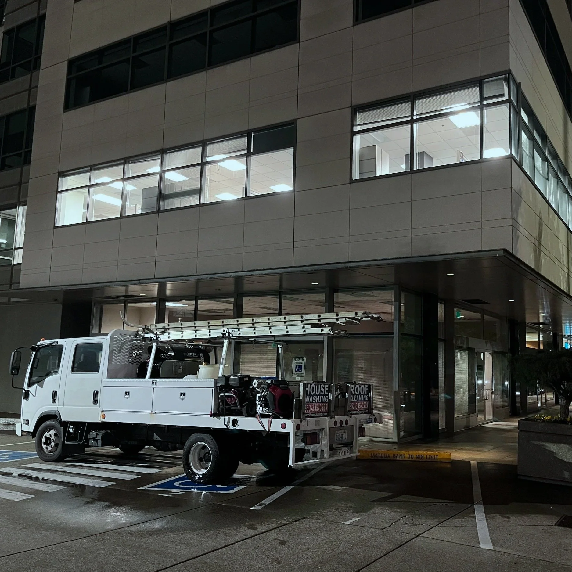 A white utility truck with a ladder on top parked outside a modern office building at night.