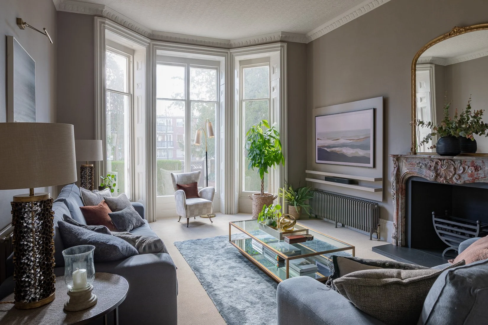 English townhouse living room with slate blue linen sofas and various textured cushions, large bay windows, an occasional armchair and neutral-colored walls.