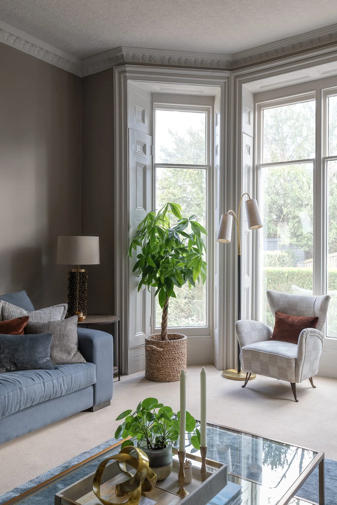English townhouse living room with slate blue linen sofas and various textured cushions, large bay windows, an occasional armchair and neutral-colored walls.