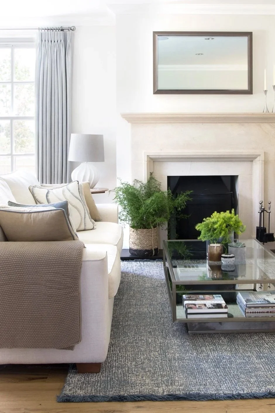 Bright and airy living room with ivory couches, a glass coffee table, a fireplace, decorative cushions, a wall mirror, and artwork.