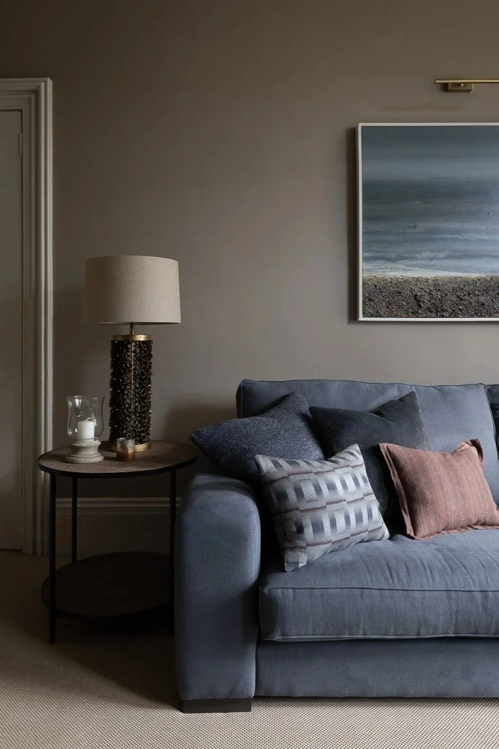 A moody living room with slate blue linen sofa with various textured pillows, bronze table lamp,  a wall with an abstract landscape painting above the sofa, and neutral-colored walls.