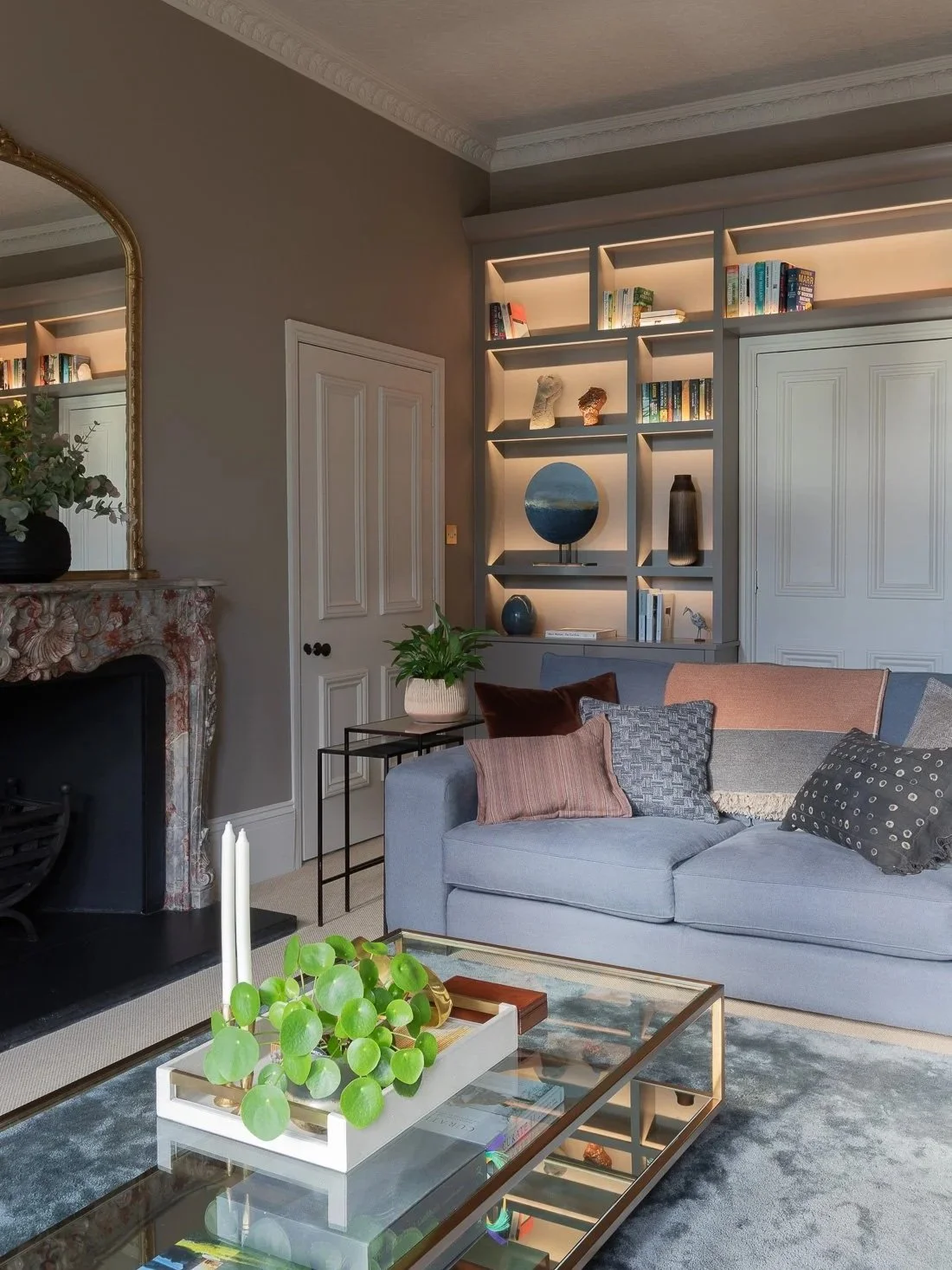 A living room with slate blue linen sofas and various textured cushions, bespoke built-in bookcase shelving and storage with integrated lighting, neutral walls and partition double doors.
