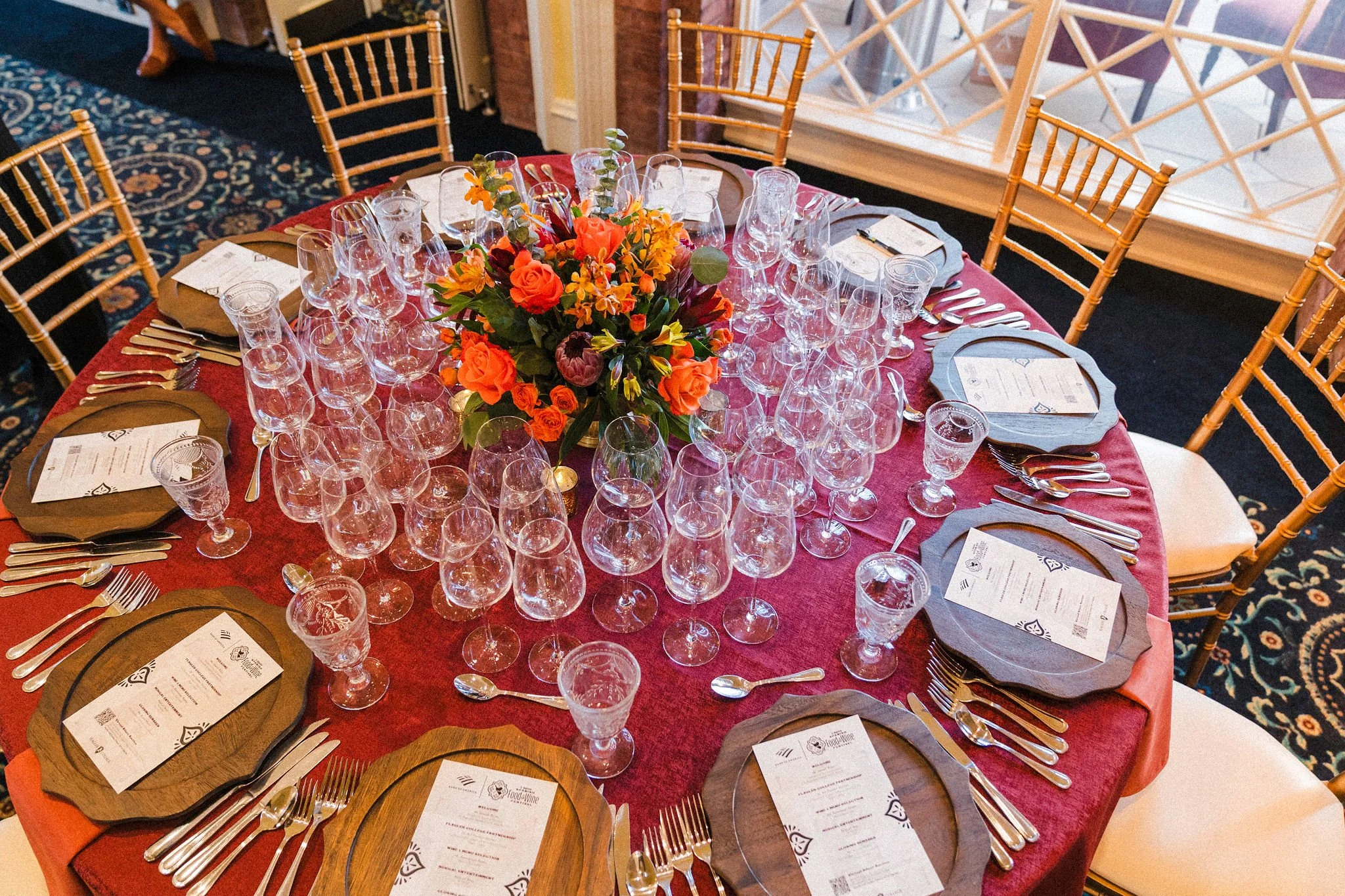 Elegant dining table setup with floral centerpiece, gold plates, glassware, and menus on patterned tablecloth at an event.