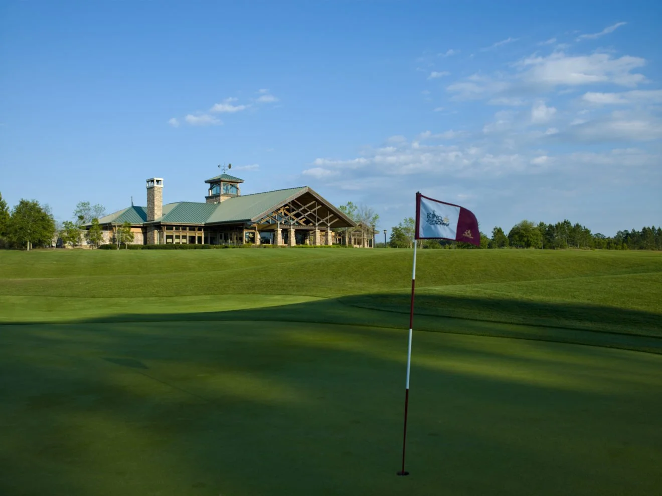 Golf course with a green, flagstick, and clubhouse in the background under a blue sky.