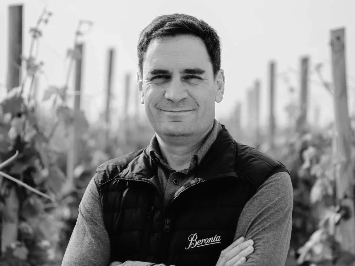 A man with short dark hair, wearing a vest with "Beronia" embroidered on it, standing outdoors among grapevines, smiling with arms crossed.