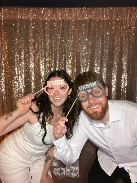 A smiling woman and man at a celebration, holding playful photo booth props that say "WIFE" and "GROOM," with a gold sequin backdrop.