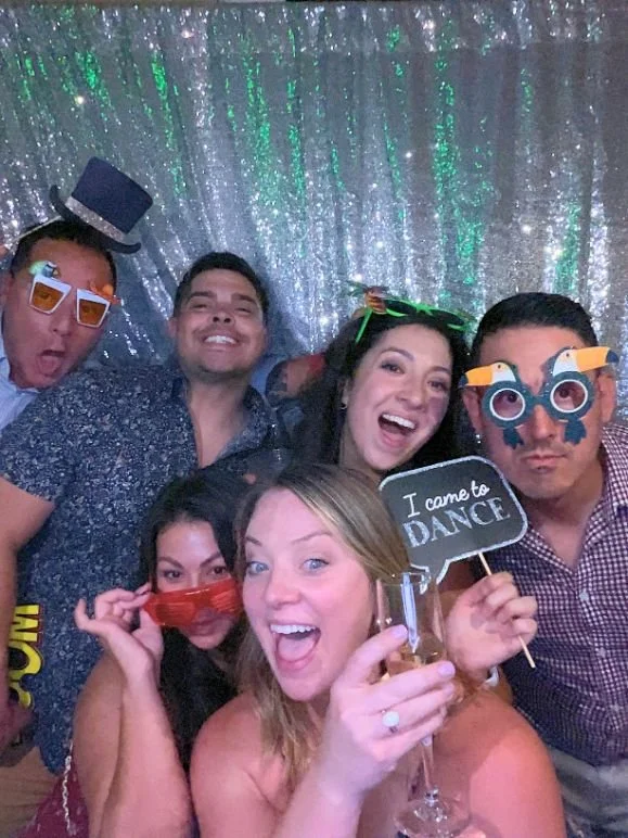 A group of six people at a party, wearing playful costumes and props, posing in front of a silver, glittery backdrop, with one person holding a speech bubble sign that says "I came to dance," and another woman holding a drink.