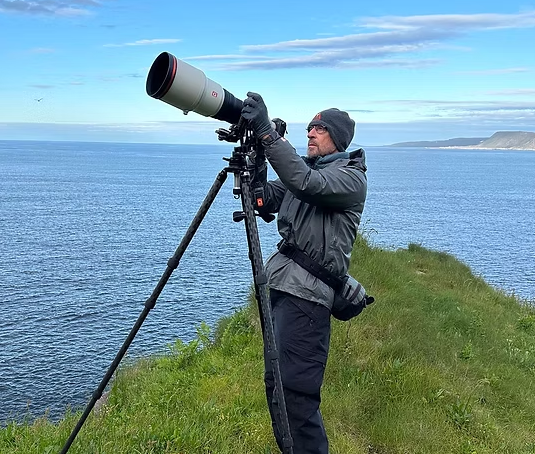 Wildlife photographer, Peter Cavanagh, using a telephoto camera lens on a tripod atop a grassy coastal cliff overlooking the ocean