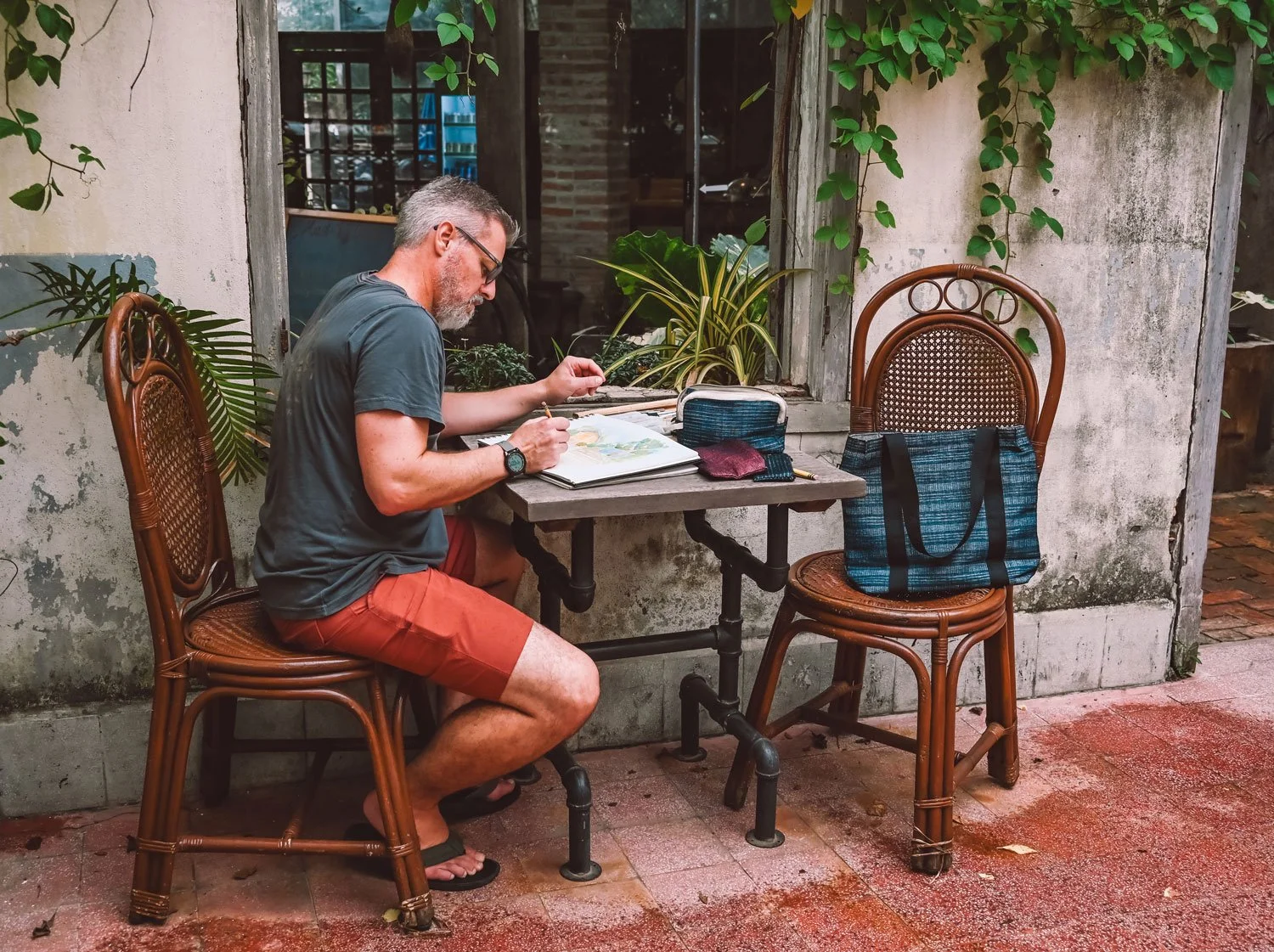 A man with gray hair, glasses, and a beard is sitting outdoors at a small table, drawing or painting in a sketchbook. He is wearing a dark t-shirt, orange shorts, and flip-flops. There are two wicker chairs around the table, one unoccupied with a blu