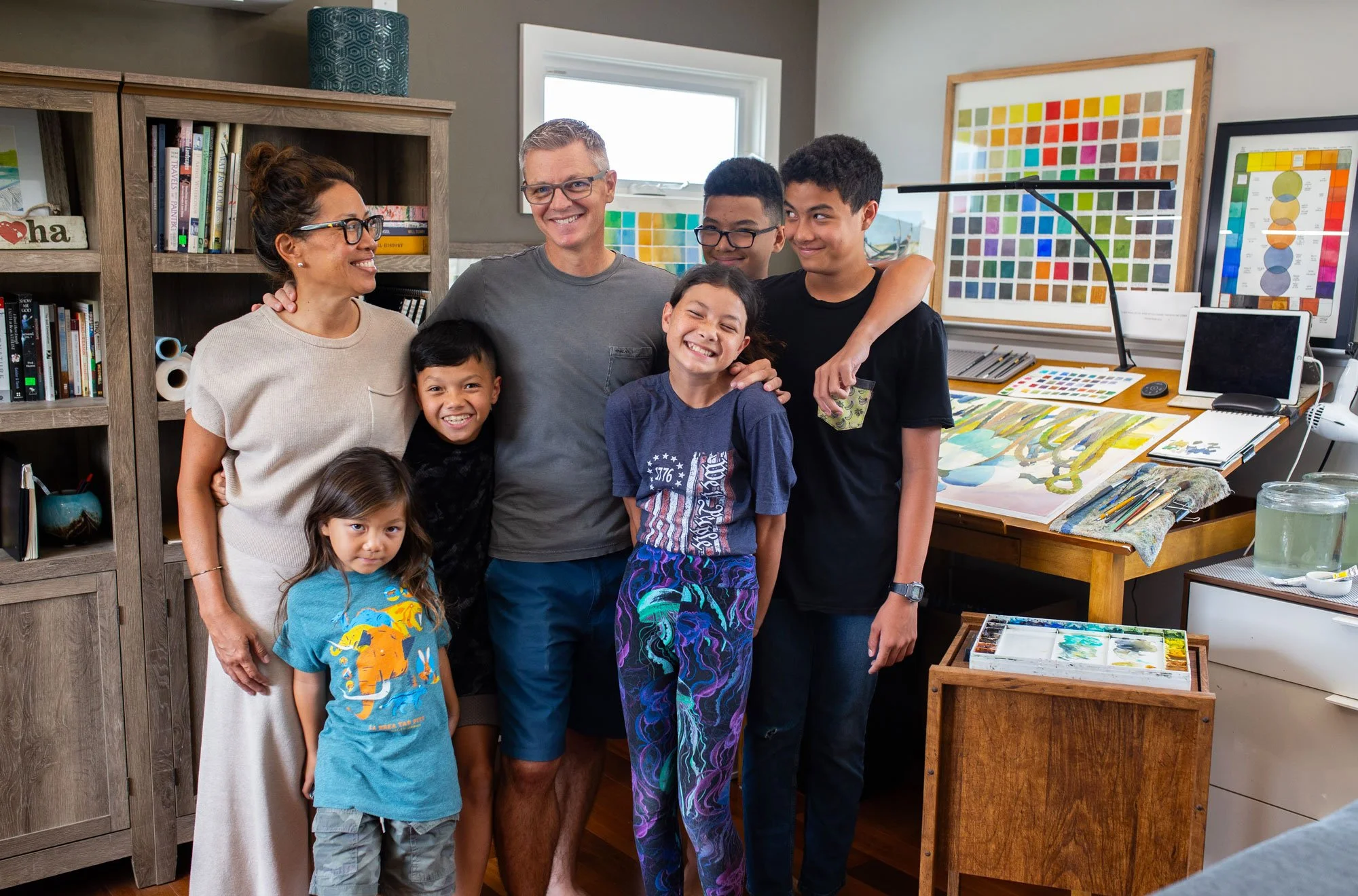 Group of seven people, including children and adults, gathered in an art studio, smiling and celebrating. There are colorful paintings and art supplies on the tables and walls behind them.