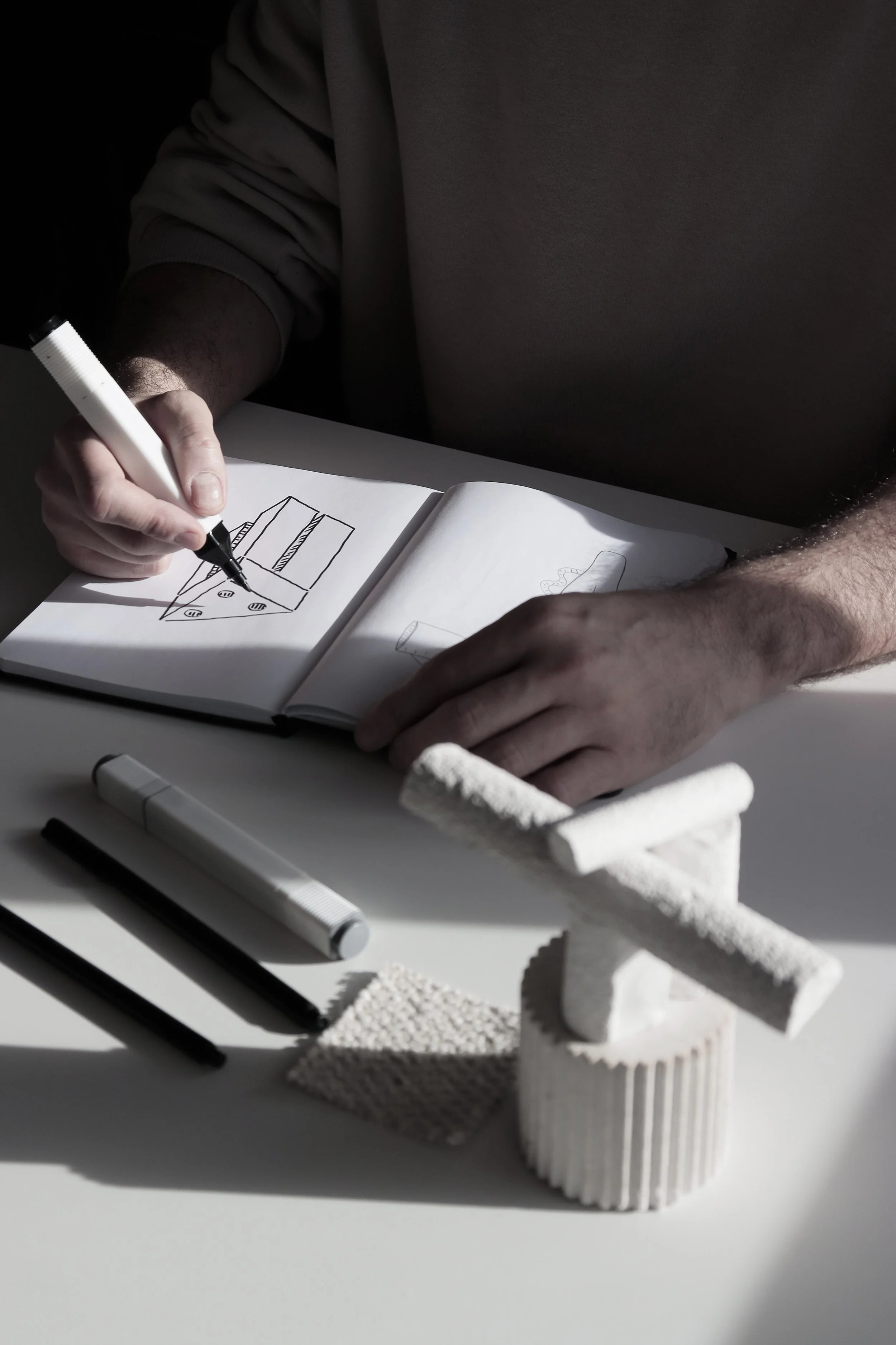 Persona dibujando en un cuaderno con varios marcadores en una mesa que tiene objetos decorativos blancos.