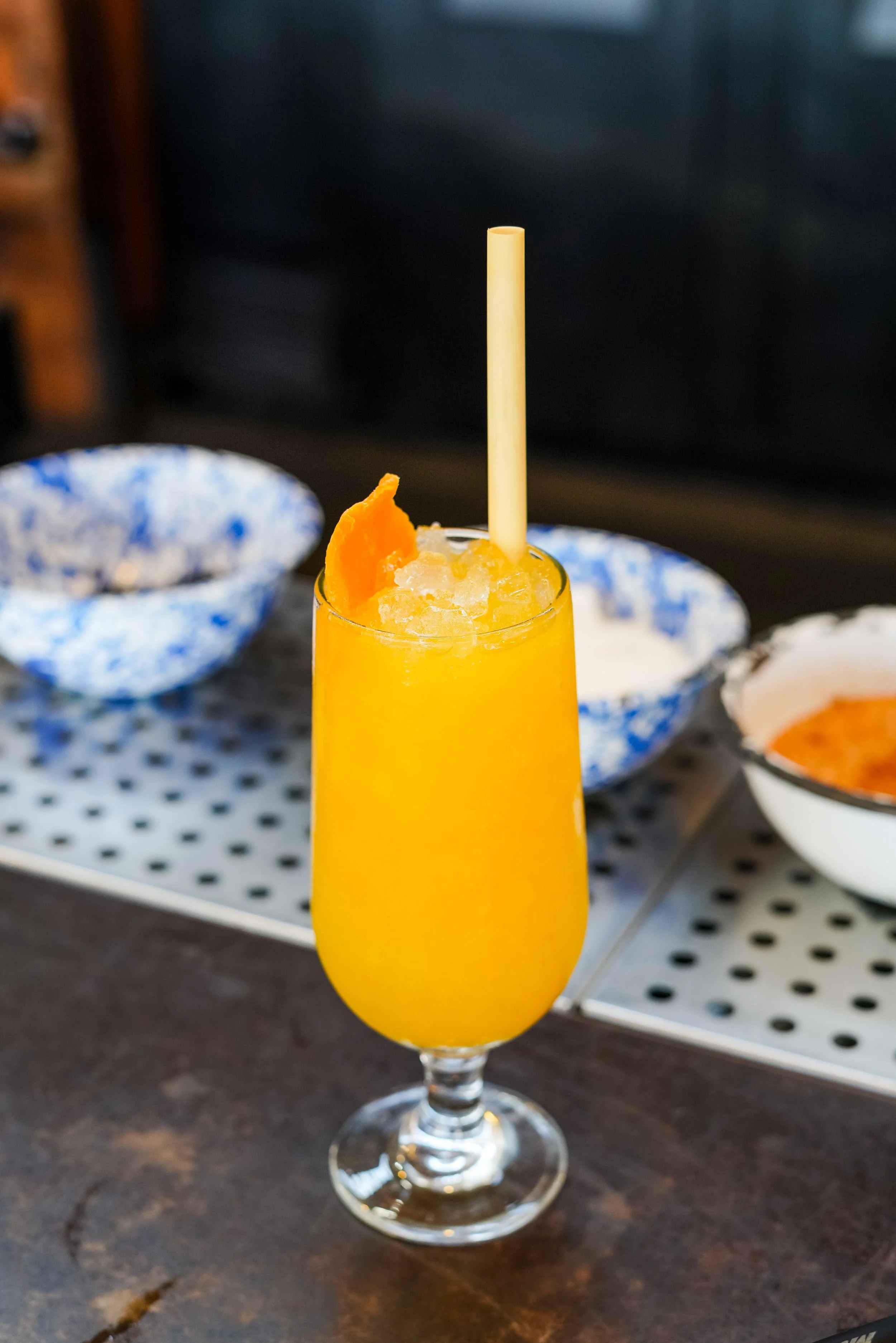 A tall glass of orange mango juice garnished with a mango slice on the rim, with a white straw, on a dark wooden table with bowls of ingredients in the background.
