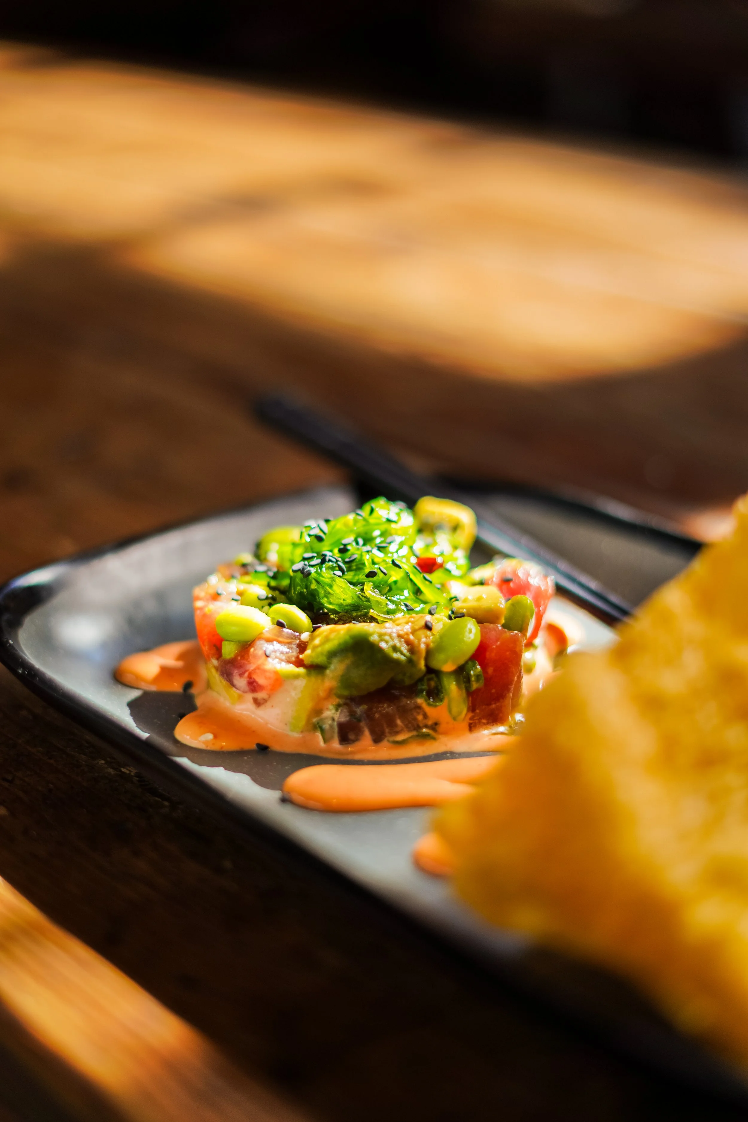 Close-up of a colorful appetizer with avocado, tomato, and green onions on a black ceramic plate with a drizzle of orange sauce.