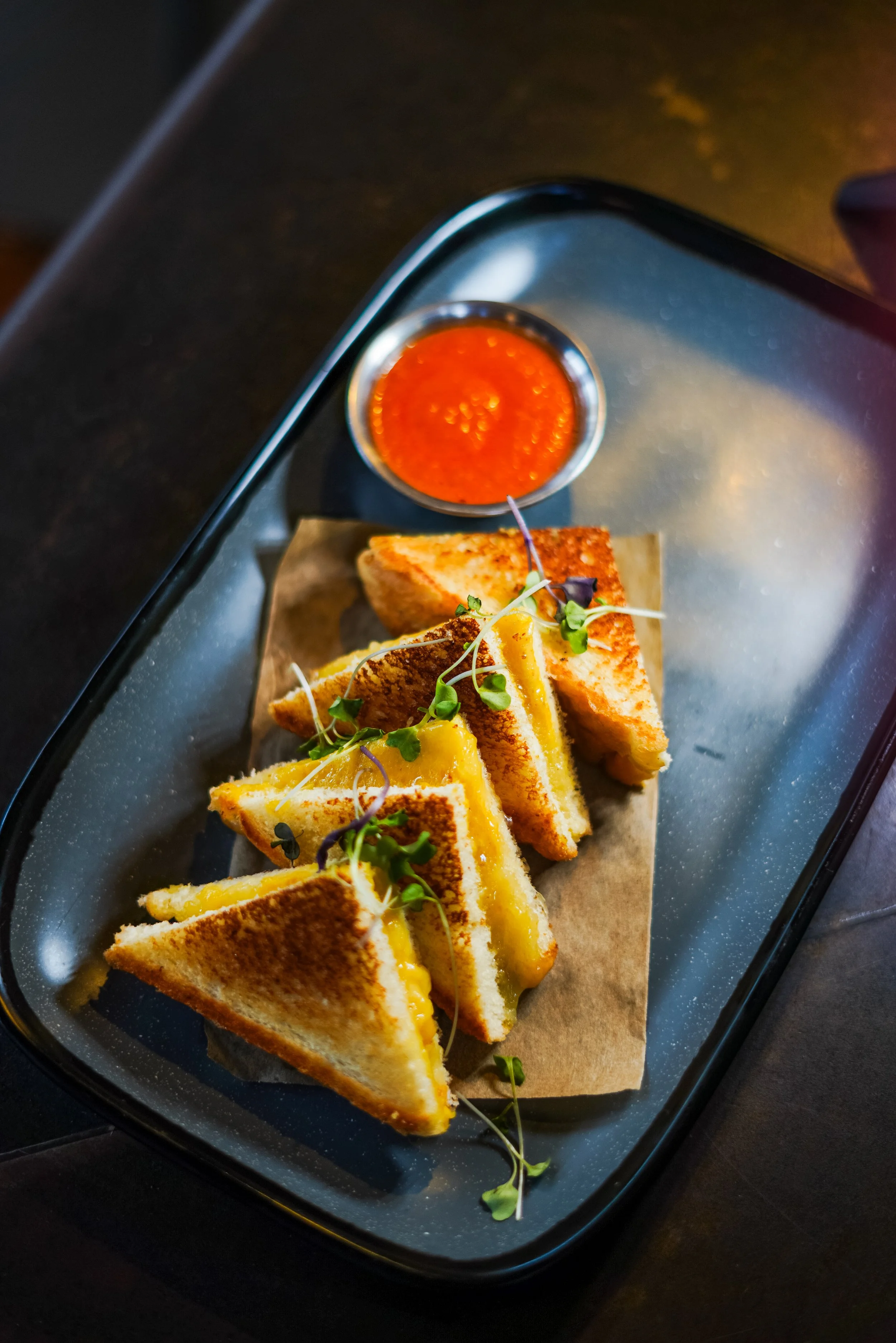 Close-up of grilled cheese sandwiches cut into triangles, served with a small bowl of red dipping sauce, garnished with microgreens, on a black serving tray.