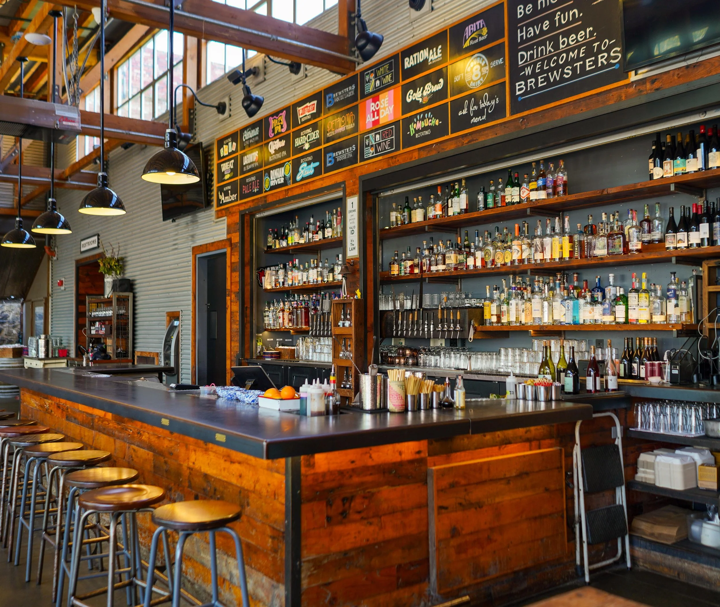 Inside a bar with a high wooden counter and bar stools. Shelves behind the bar are stocked with various bottles of alcohol. Signage above displays drink options and a welcome message.