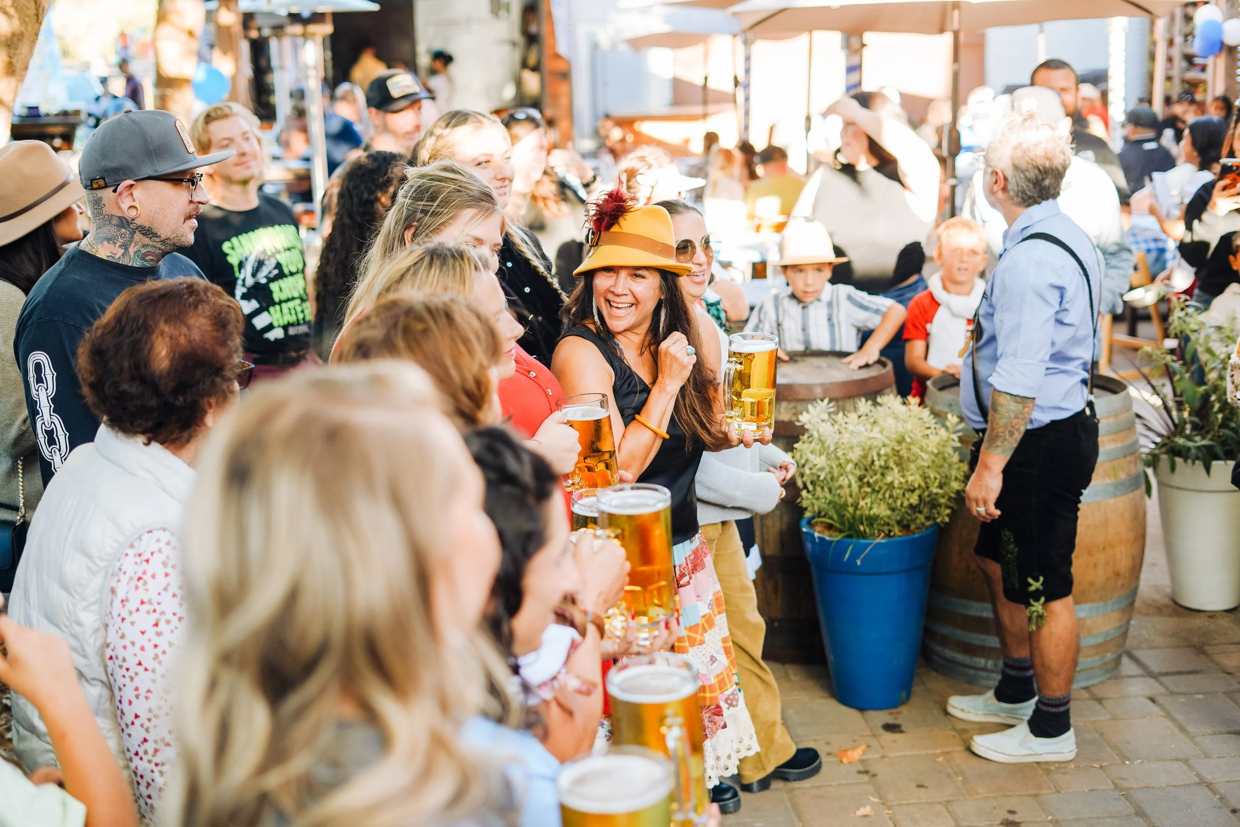 People at an outdoor event, some holding glasses of beer, engaged in conversations and smiling, with vendors and decorations in the background.