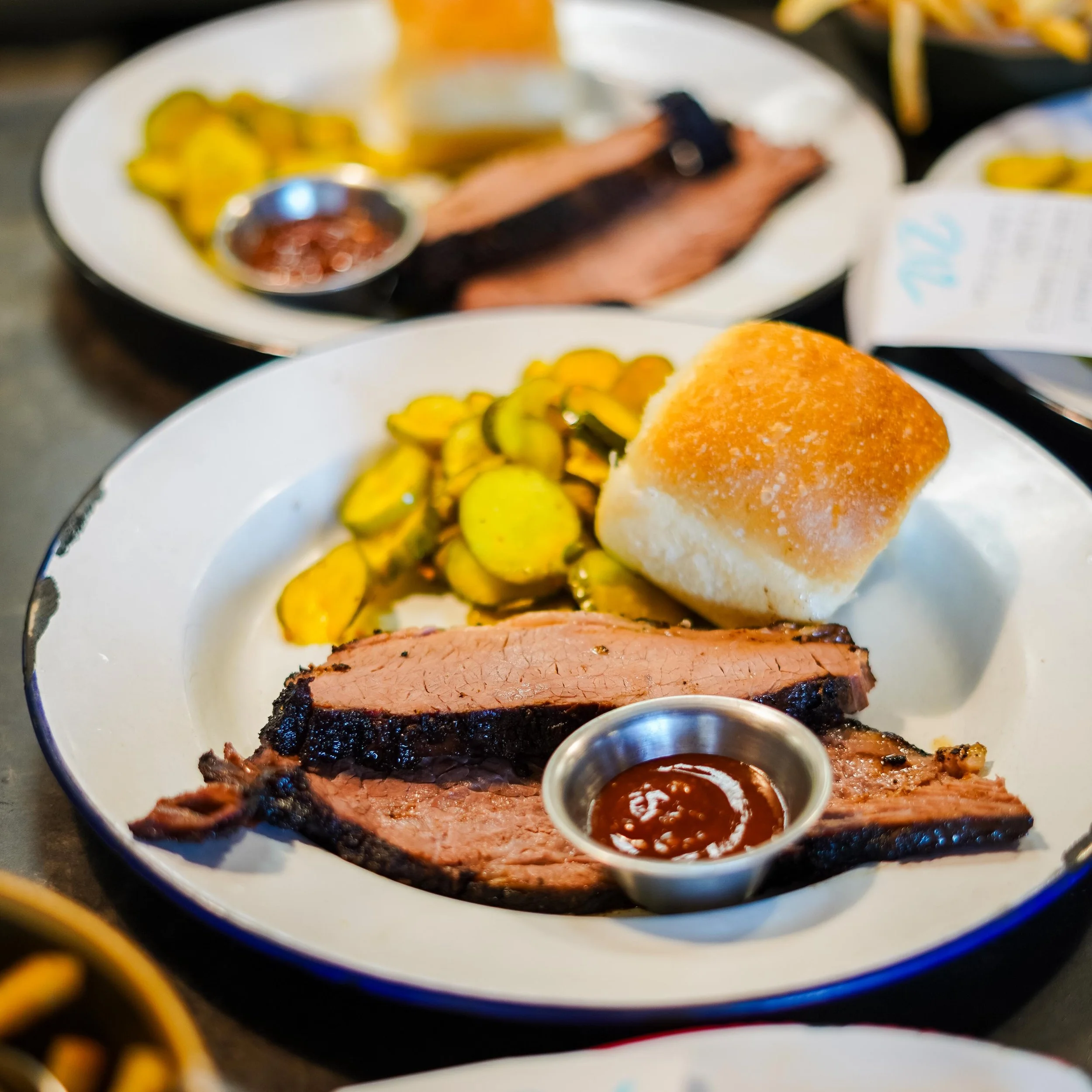 Close-up of a plate with sliced pickles, a baked potato with butter, and sliced meat with a piece of barbeque brisket, accompanied by small containers of barbecue sauce.