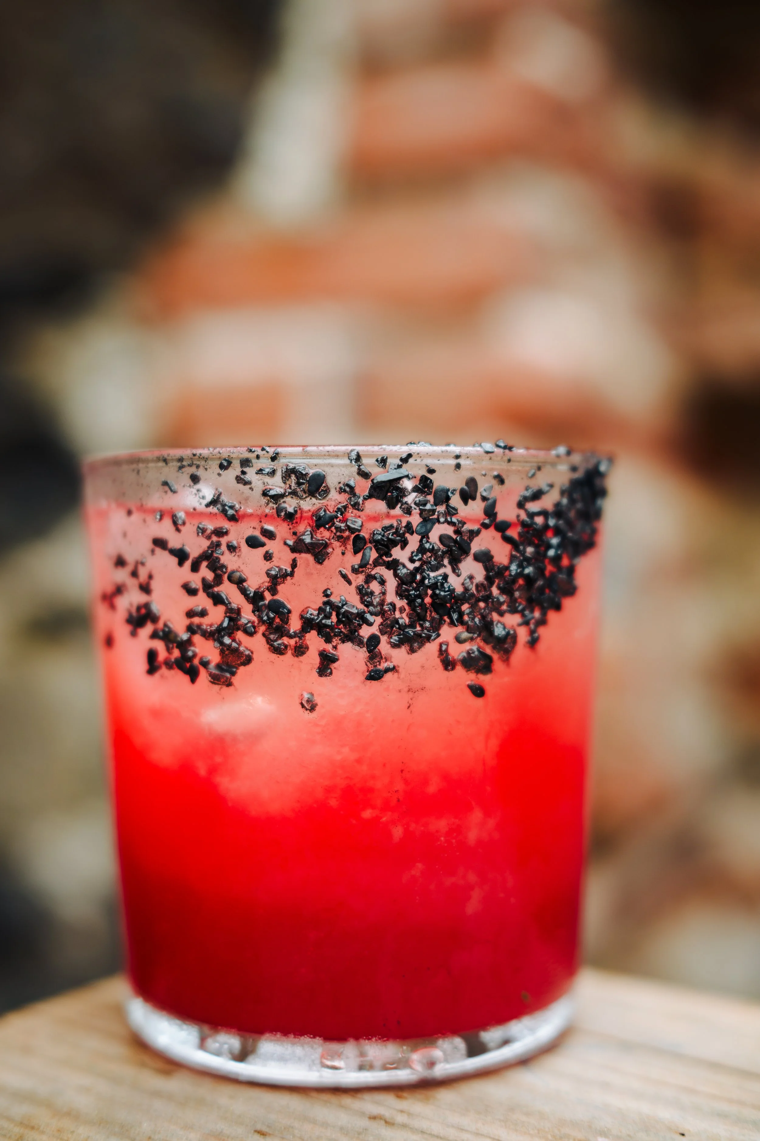 A red cocktail in a glass with black sprinkles on the rim, placed on a wooden surface with a blurred background.