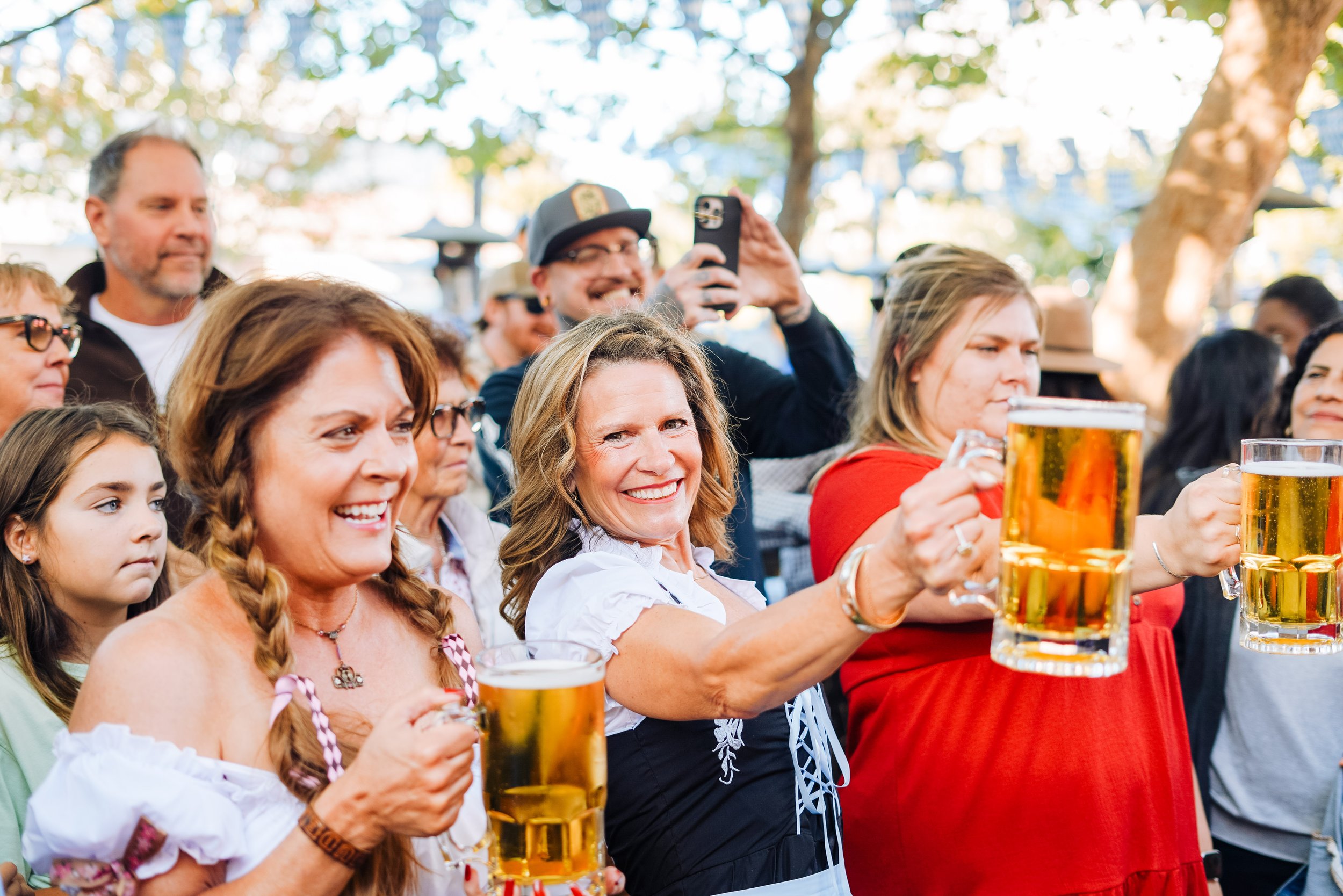 A group of people celebrating outdoors, some holding glasses of beer, with trees and sunlight in the background.