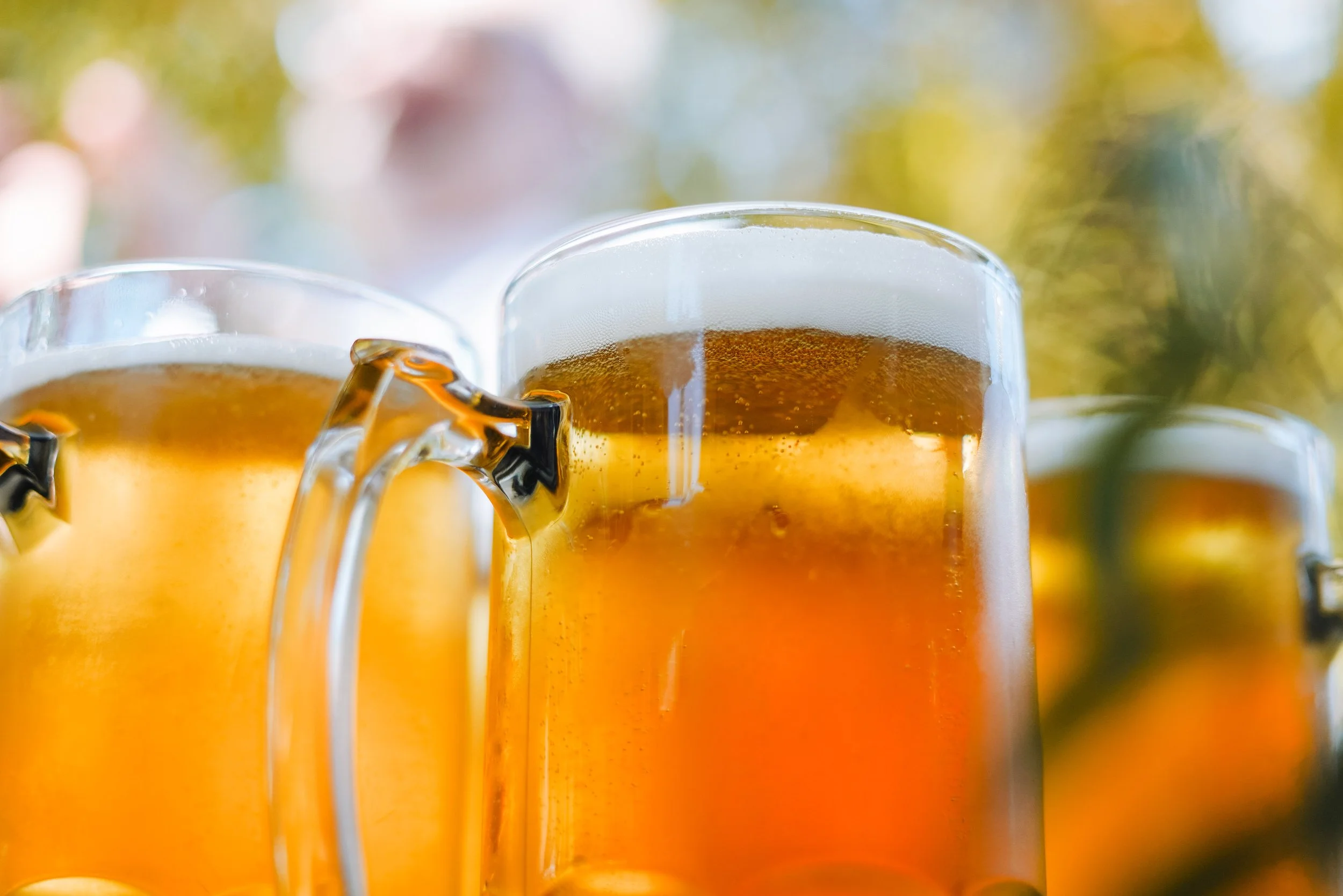 Close-up of three beer mugs filled with frothy golden beer, set outdoors with blurred green and yellow foliage in the background.