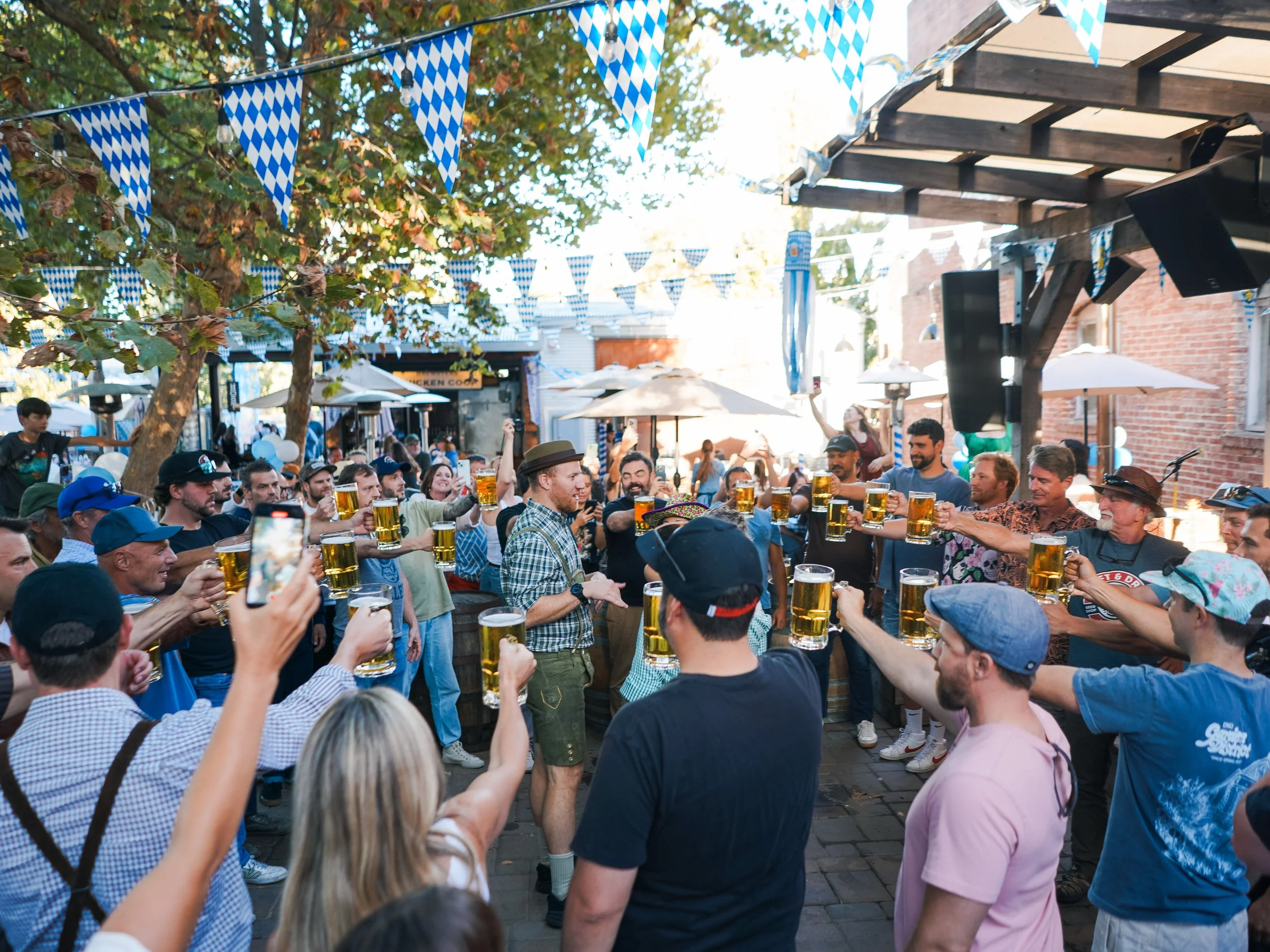 A crowd of people celebrating Oktoberfest outdoors under blue and white decorative banners, holding up glasses of beer in a toast.
