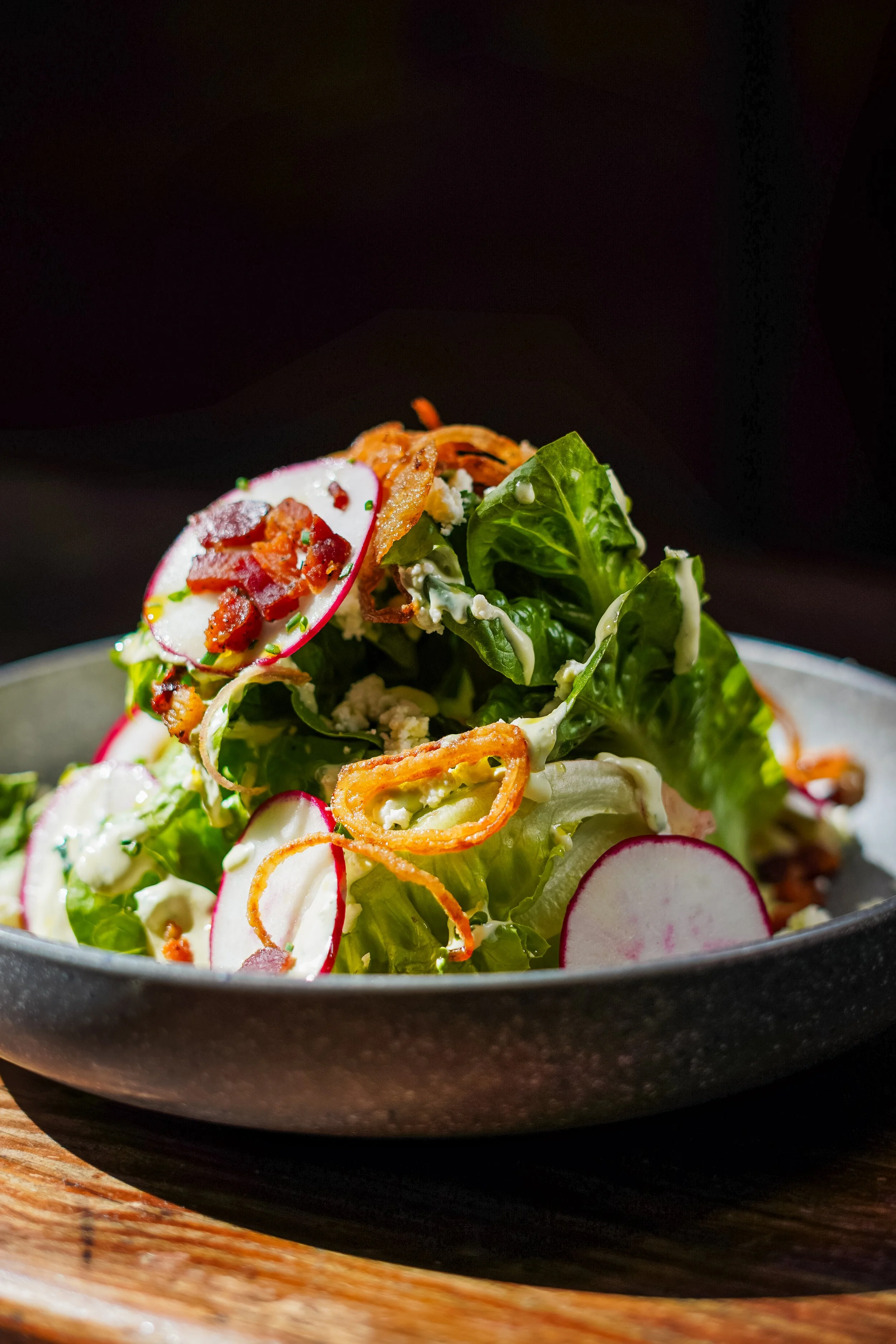 A plate of salad with pancetta,radish, feta cheese, crispy shallots, and green goddess dressing on a rustic wooden table.