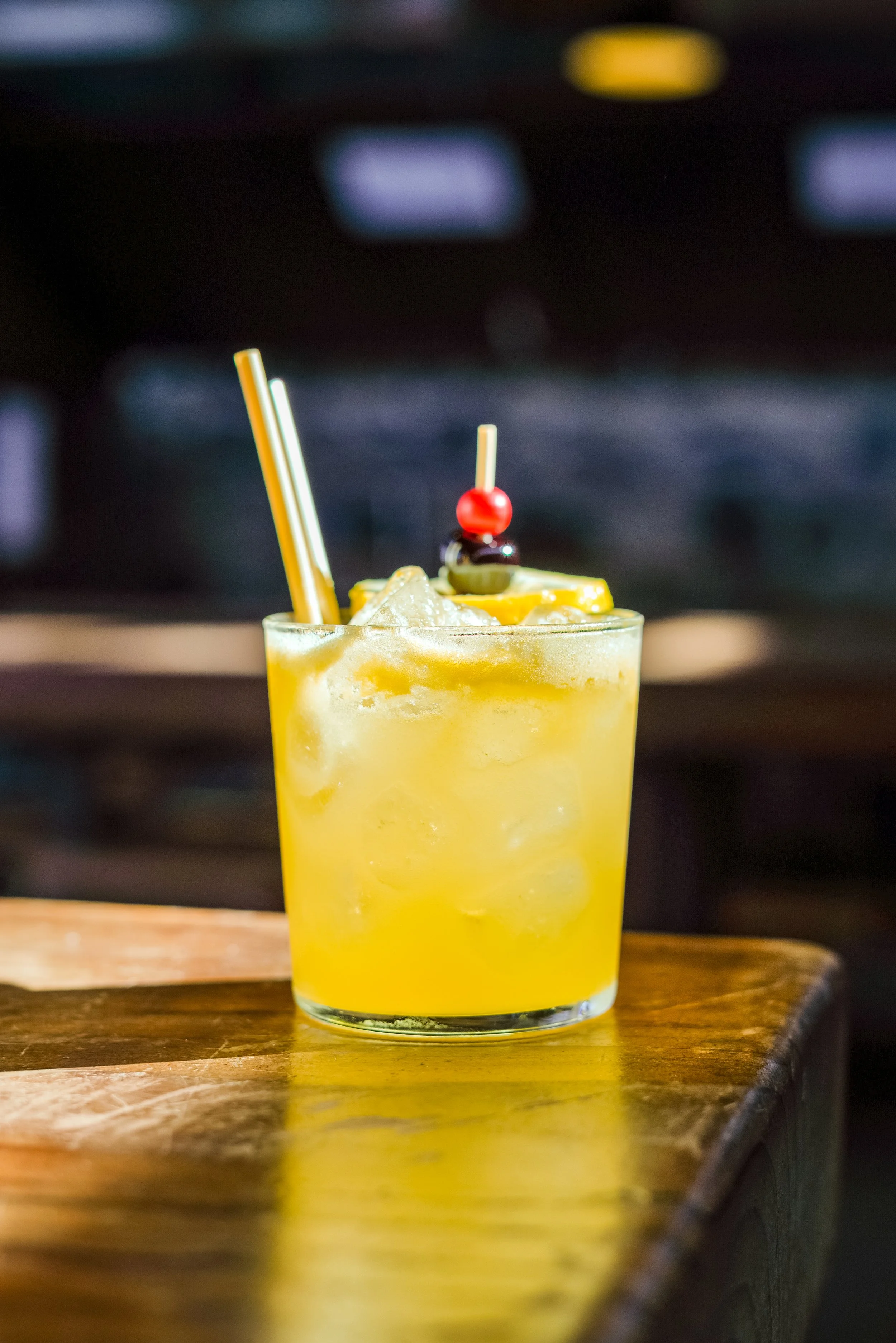 A yellow cocktail garnished with a cherry, olive, and pineapple slice on a wooden surface.