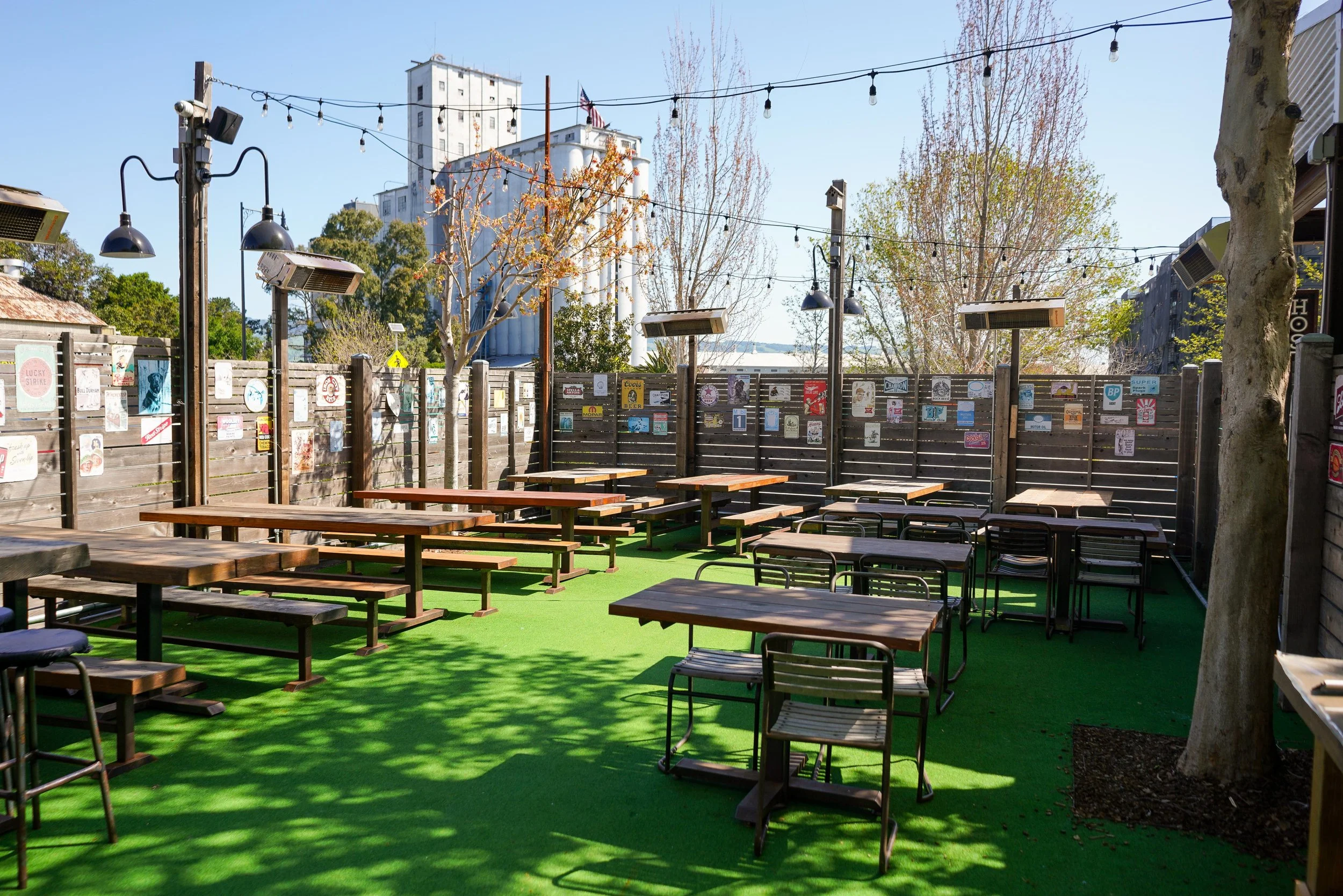 An outdoor patio with green artificial turf, wooden picnic tables, chairs, and a wooden fence decorated with various posters and art. There are string lights overhead and trees with autumn foliage.