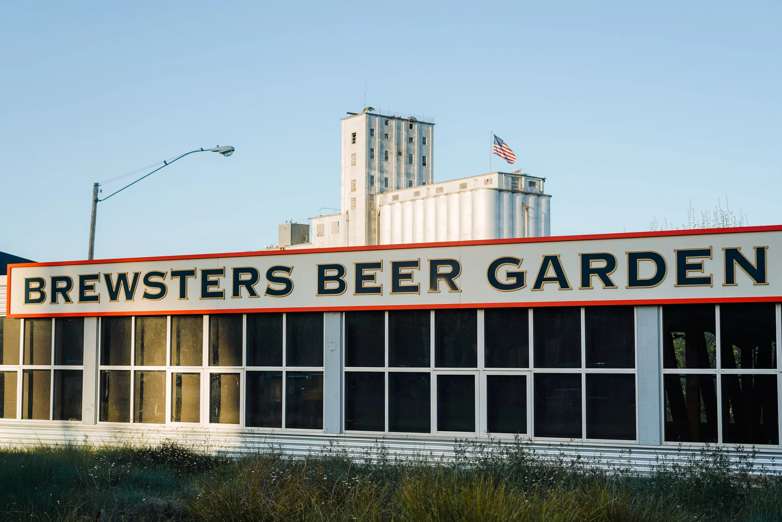 Exterior view of Brewsters Beer Garden building with large sign, windows, and an American flag on top, with a tall industrial building and a streetlight in the background.