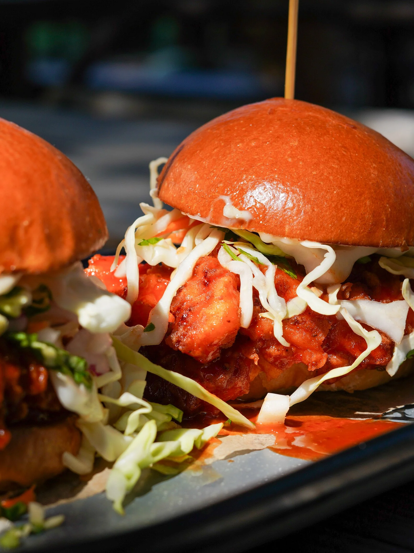 Close-up of a slider burger with a sesame seed bun, shredded lettuce, tomato, cheese, and a breaded fried chicken patty, served on a black tray.