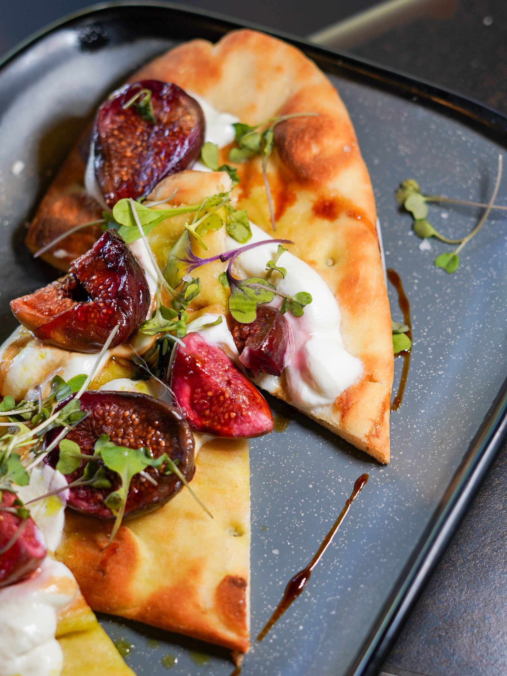 Close-up of a slice of flatbread topped with fresh figs, microgreens, dollops of cheese or sauce, and balsamic glaze on a black plate.