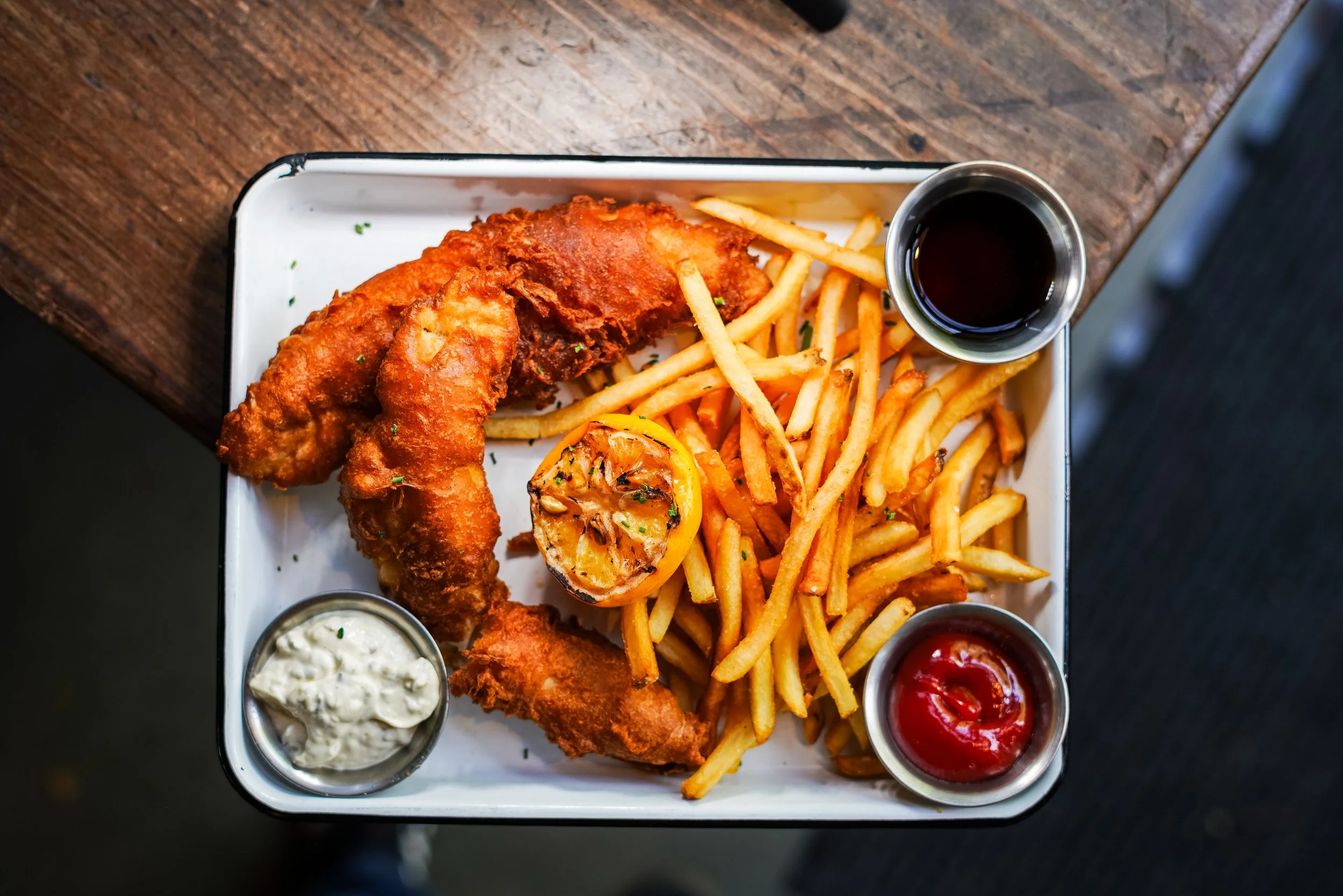 A tray with fried fish fillets, French fries, a lemon half, and three small containers of tartar sauce, ketchup, and dark sauce on a wooden table.