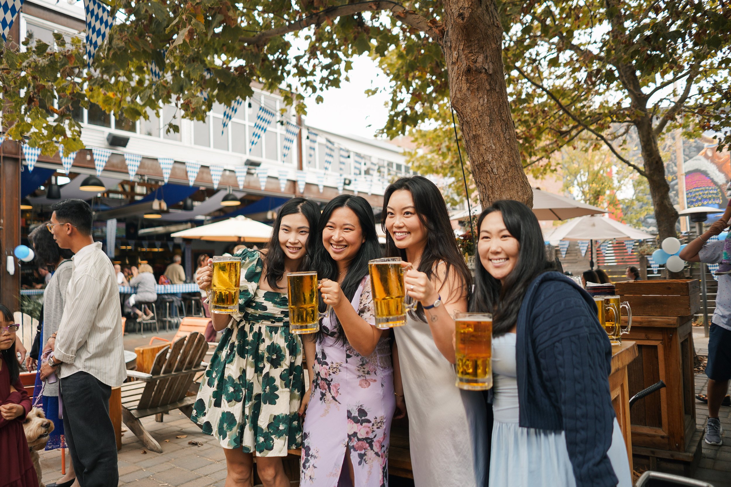 Group of women and men celebrating outdoors with beer in a festive setting decorated with blue and white banners and autumn leaves.