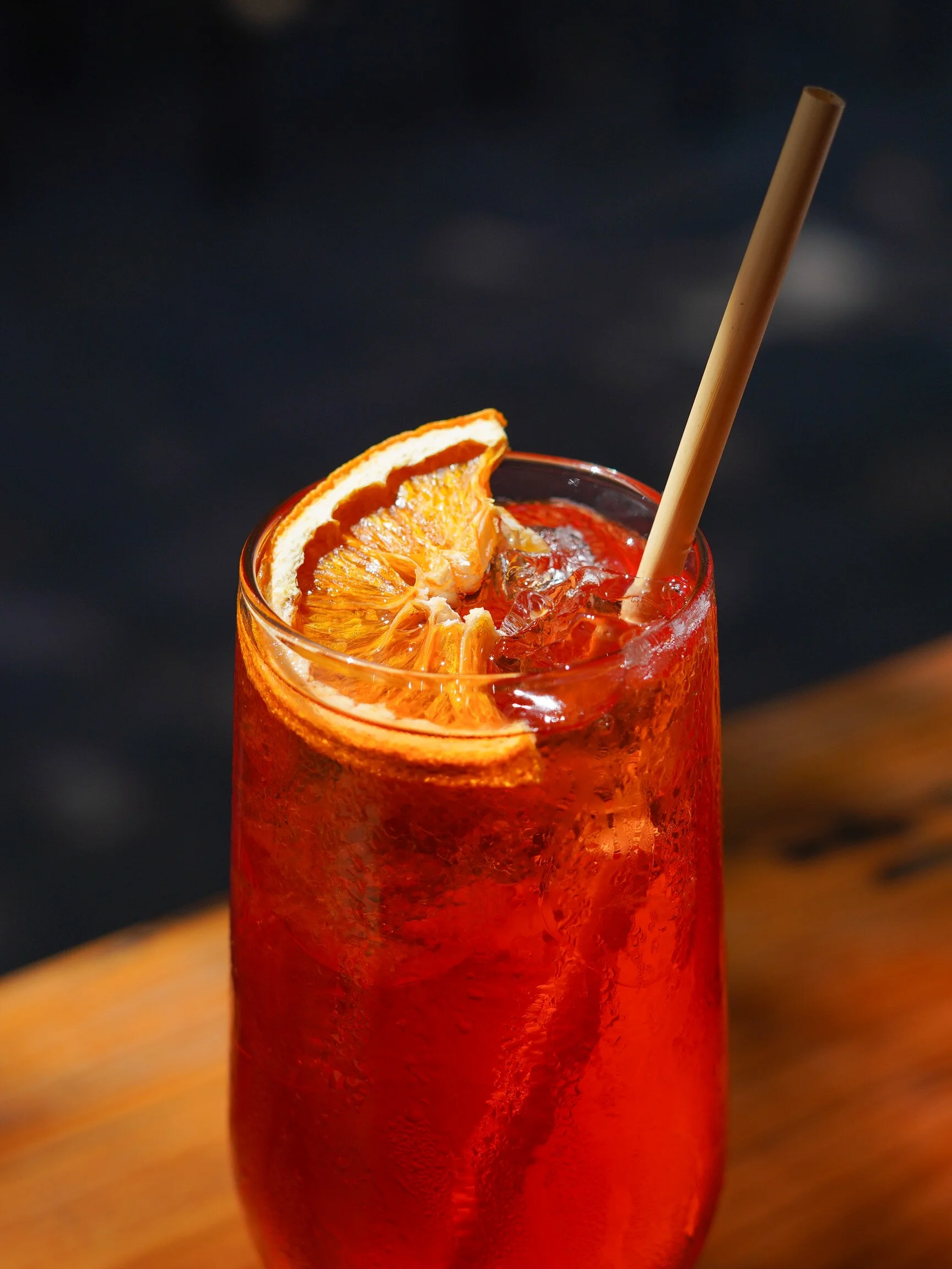 Close-up of a red cocktail drink garnished with a slice of orange and a straw, on a wooden surface with a dark background.