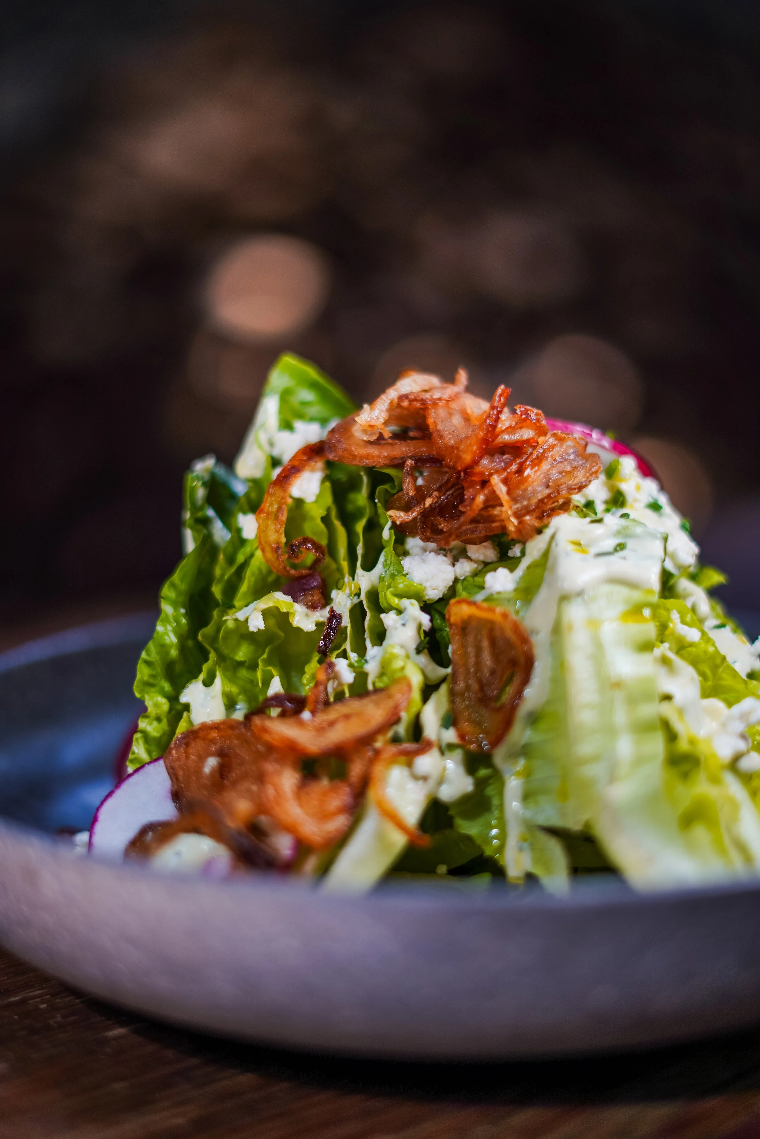 Close-up of a fresh lettuce salad with bacon, cheese, radish slices, and creamy dressing in a gray bowl.