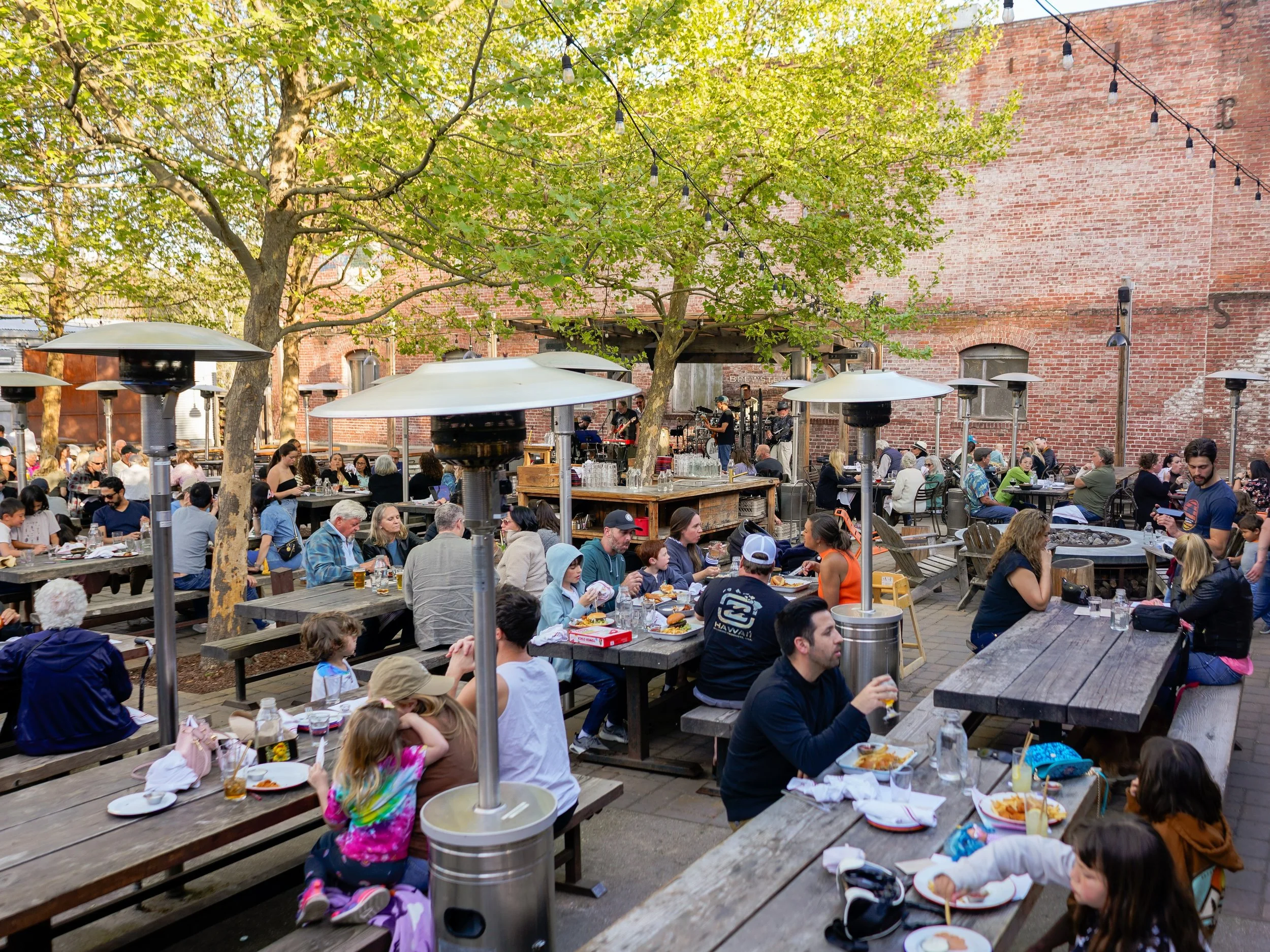 Crowd of people dining outdoors at a patio restaurant with brick walls, trees, and string lights.
