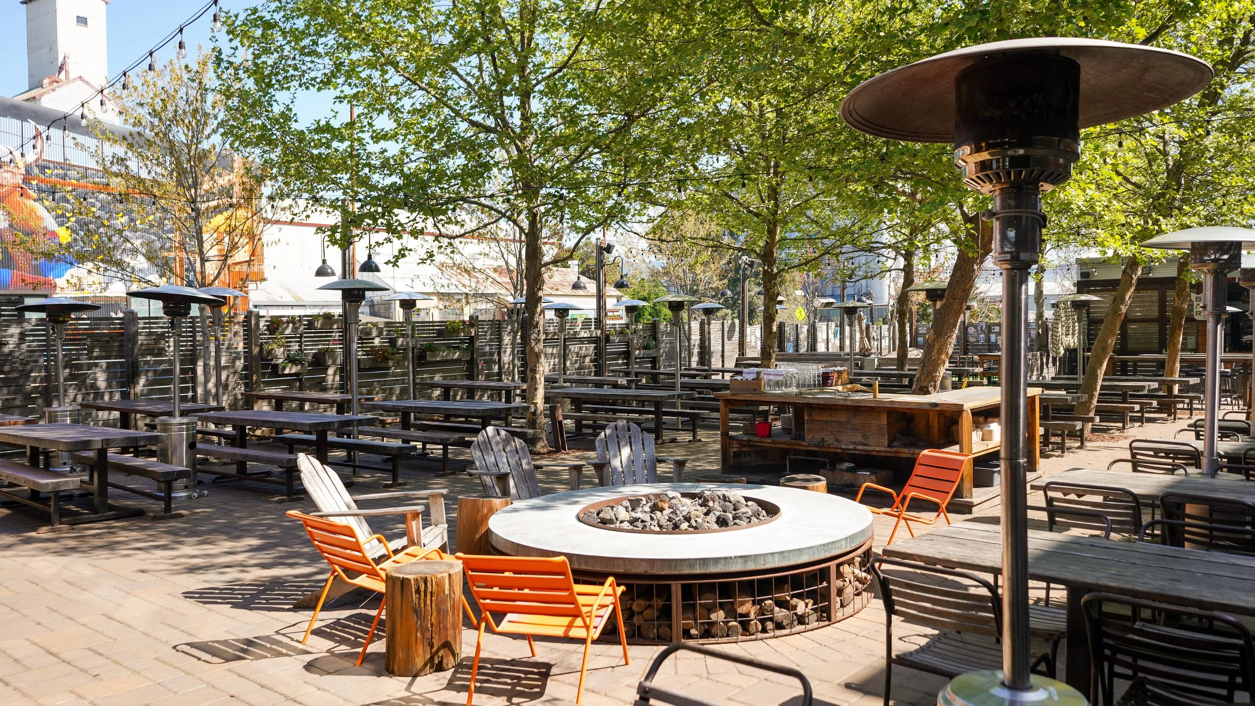 Outdoor patio with wooden tables and benches, patio heaters, a fire pit with rocks, and trees providing shade.