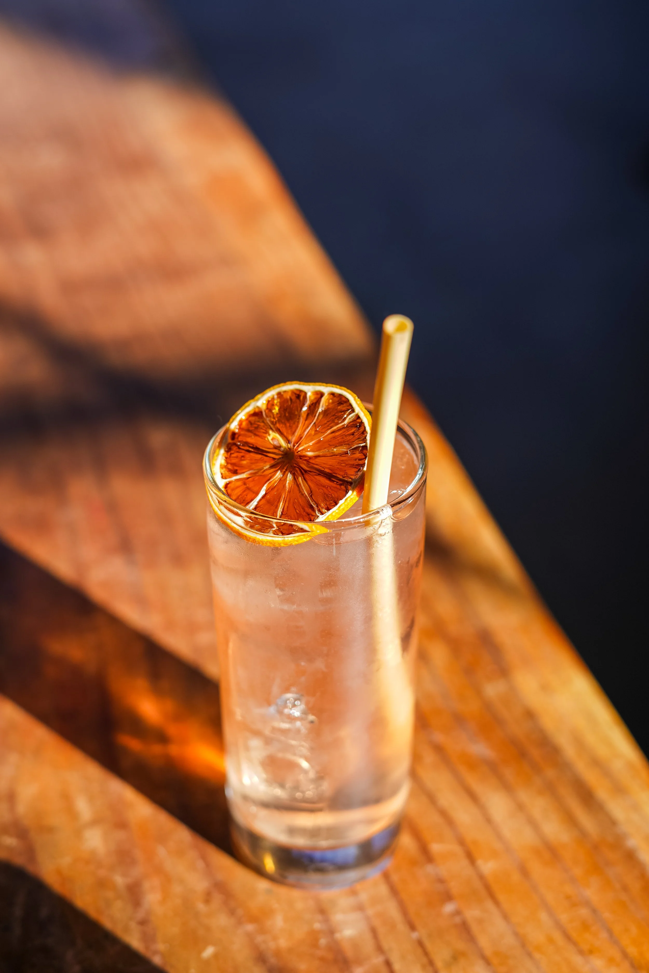 A tall glass of iced beverage garnished with a dried orange slice and a straw on a wooden surface.