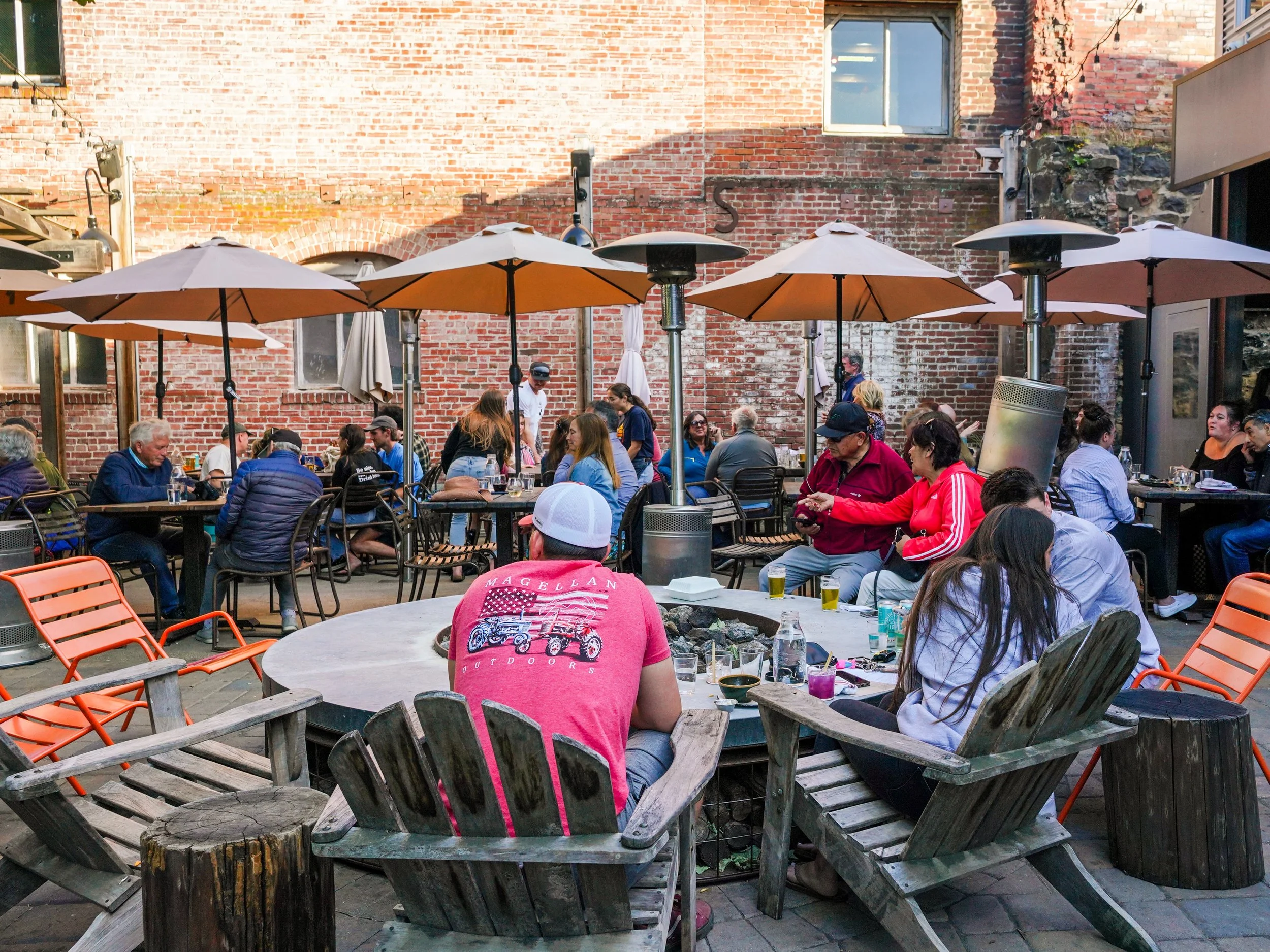 A lively outdoor gathering in a tree-shaded patio area, with string lights overhead and heaters among tables.