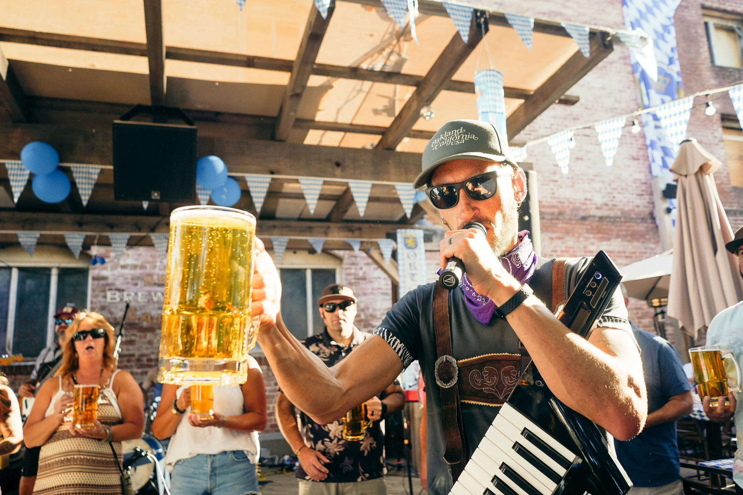 A man wearing sunglasses, a gray cap with 'Oakland, California' written on it, and a purple bandana, is holding a microphone and singing or speaking at an outdoor gathering. Around him are other people holding beers, festive bunting, and balloons, wi