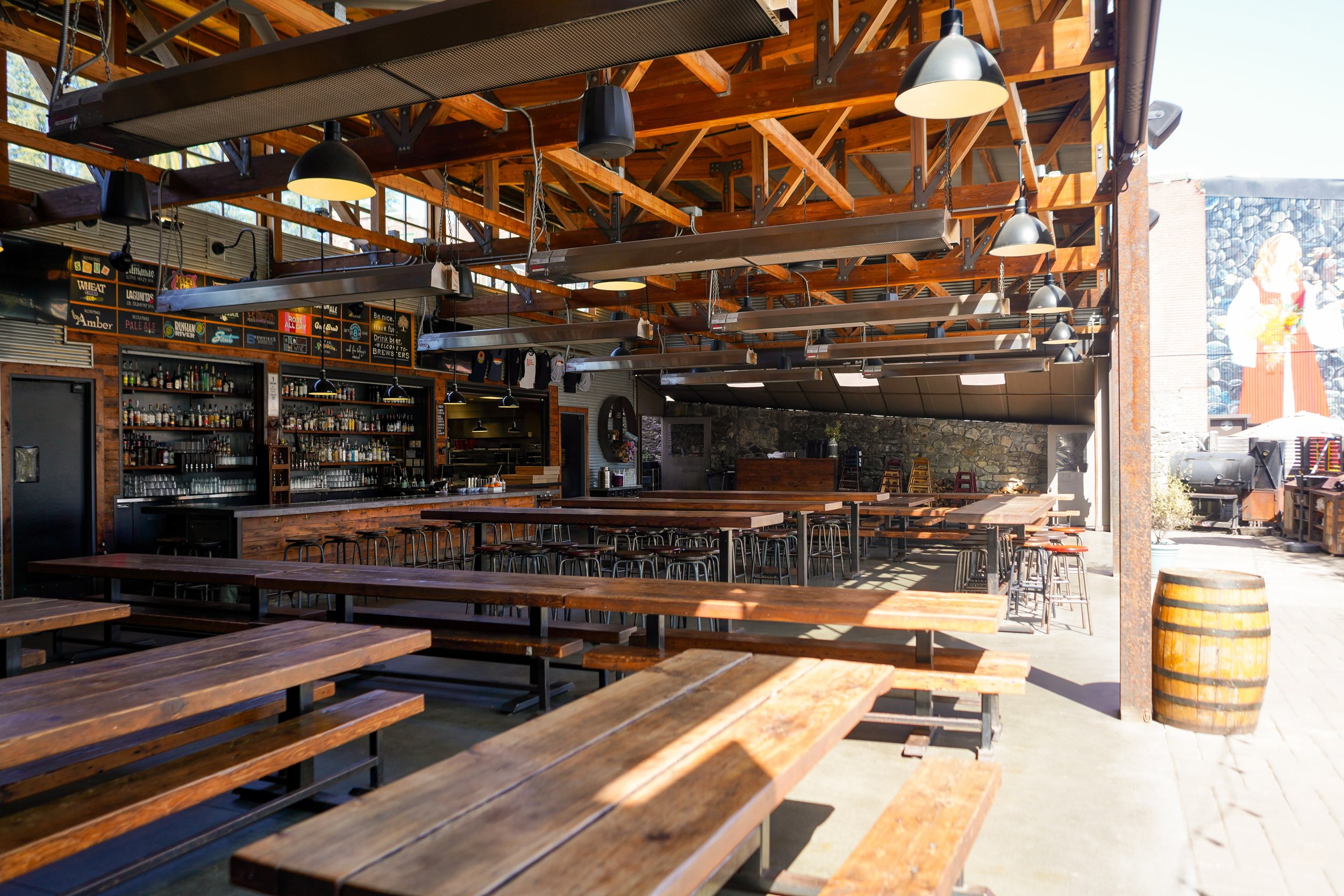 Empty indoor brewery or restaurant with wooden picnic tables and bar area, exposed wooden beams, hanging industrial lights, and a large mural on the outside wall.
