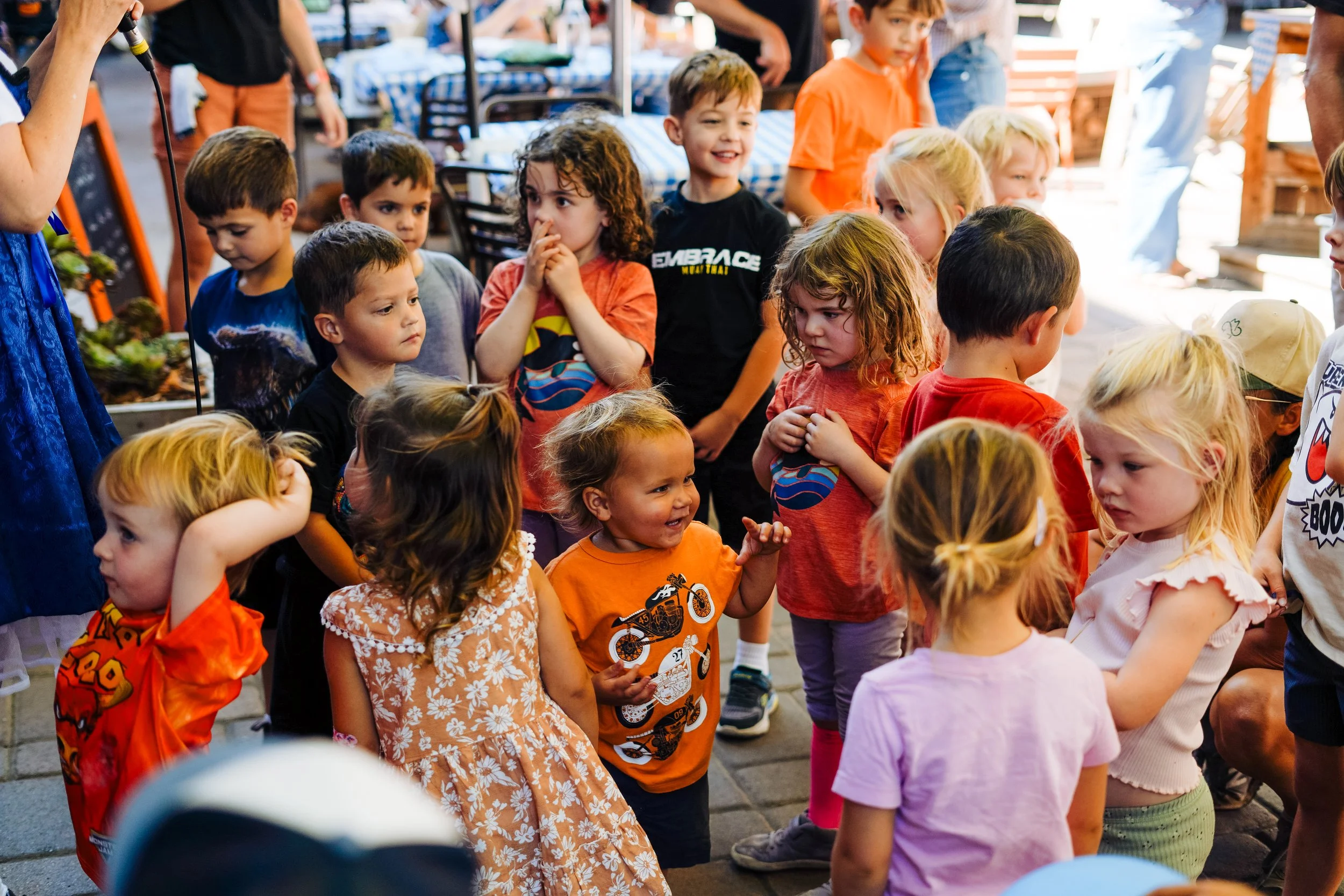 A group of children gathered outdoors, with some wearing Superman t-shirts and others in colorful clothing, participating in an activity at a daytime event.