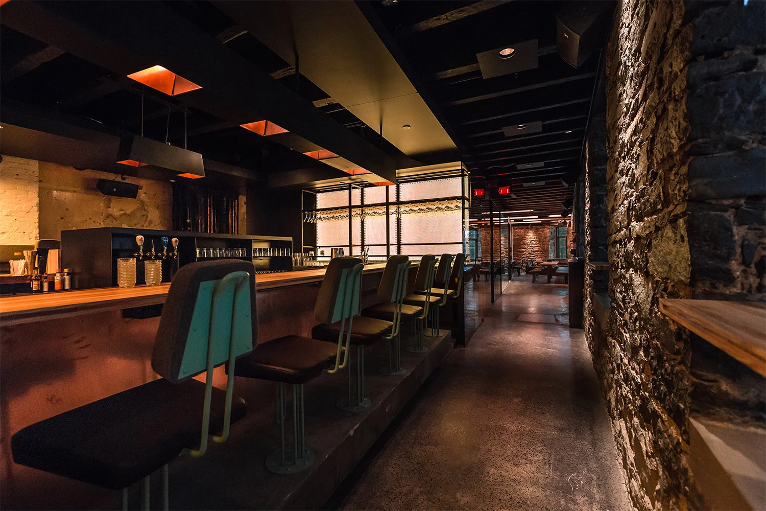 View of the bar with original stone walls and polished concrete floor. The counter, made of beech wood, surrounded by custom stools. The back of the backlit bar showcases the bottles
