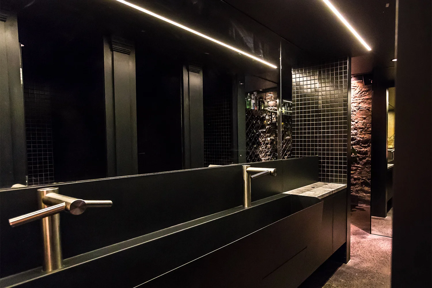 Black commercial bathroom. Automatic faucets installed on the washbasin made to measure in black finish metal. A large mirror with a black mosaic section gives us a glimpse of the cabins