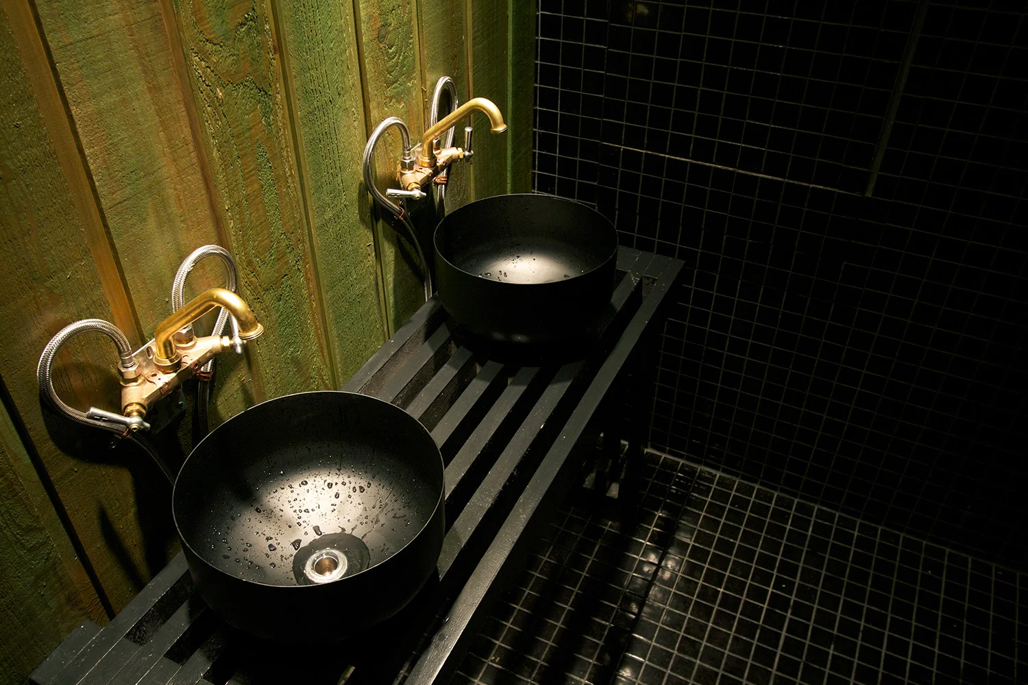 2 lavabos de salle de bain de type vasque déposés sur un comptoir de lattes de bois ajourés peint noir. La robinetterie murale brute avec arrivée d’eau visible. Mur et plancher finis mosaïque noir