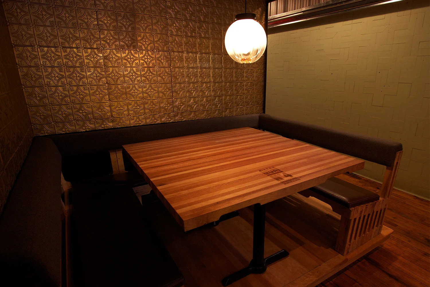 Vue d’une banquette en U fait de bois brut avec assise en cuir. Table à l’image d’une palette de transport. L’un des murs est recouvert d’anciennes tuiles de métal embossées avec motif de rosace