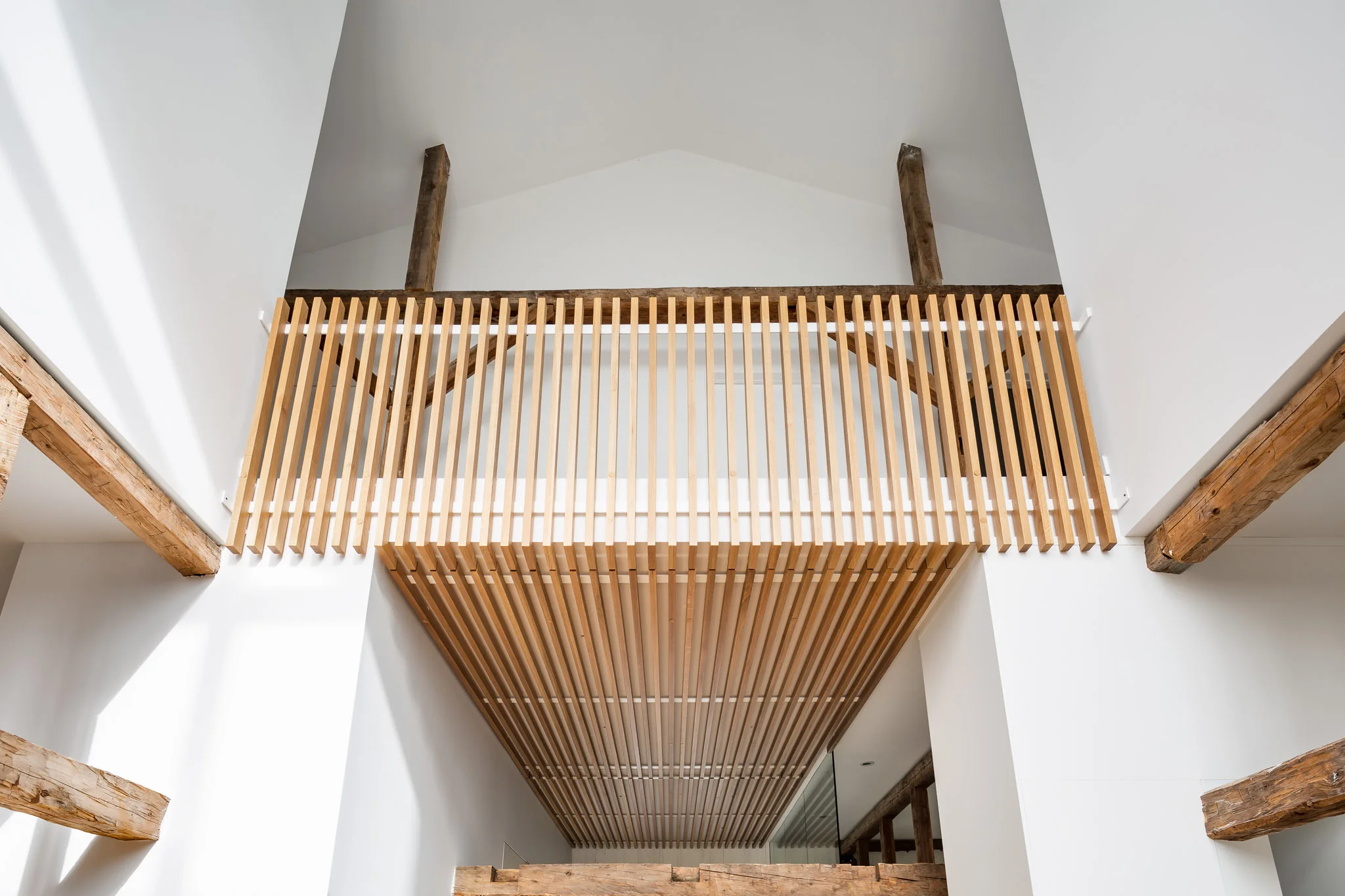 Railing in white oak slats continuing on the ceiling of the entrance. Old barn structure in apparent wood. Double height cathedral ceiling