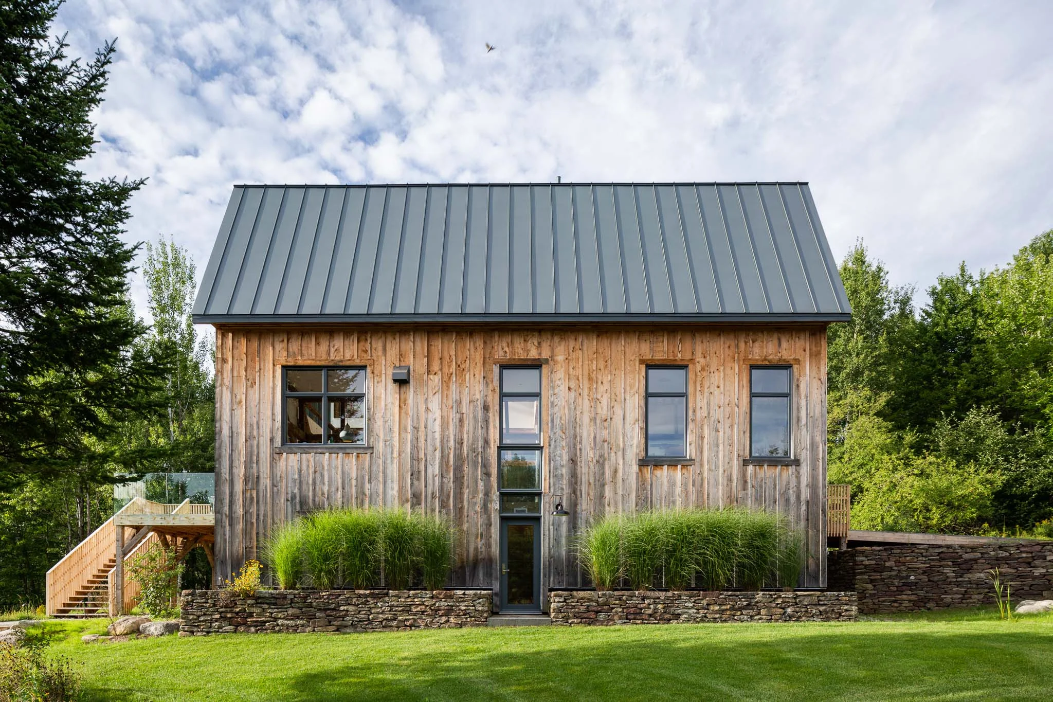Renovation of a cottage in the Eastern Townships inspired by a barn. Footbridge leading to the main entrance. Exterior cladding of boards of cedar boards and batten. Cedar deck