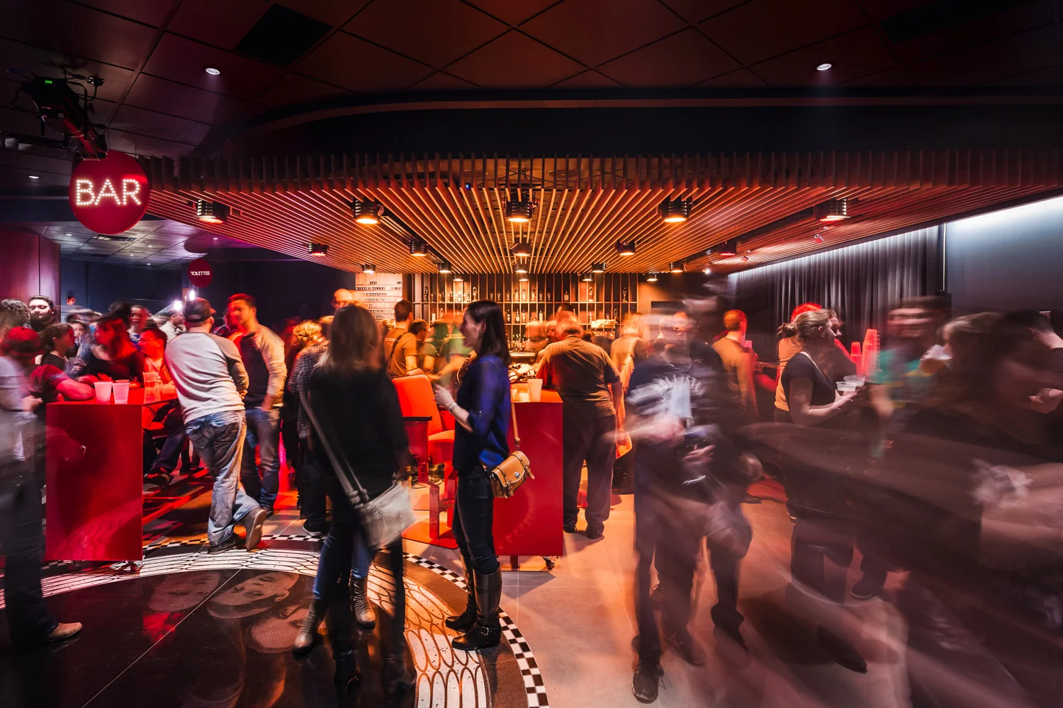 Bar avec retombée de plafond fait de lattes en noyer noir verticales et luminaires scéniques intégrés. Cercle signalétique rouge avec "BAR" en points lumineux. Mobilier rouge et clients en mouvement