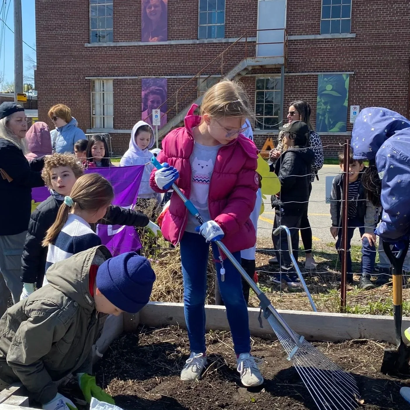 Children and adults participate in a community gardening event outside, some planting or tending to the soil, others observing or assisting, with a brick building and banners in the background.
